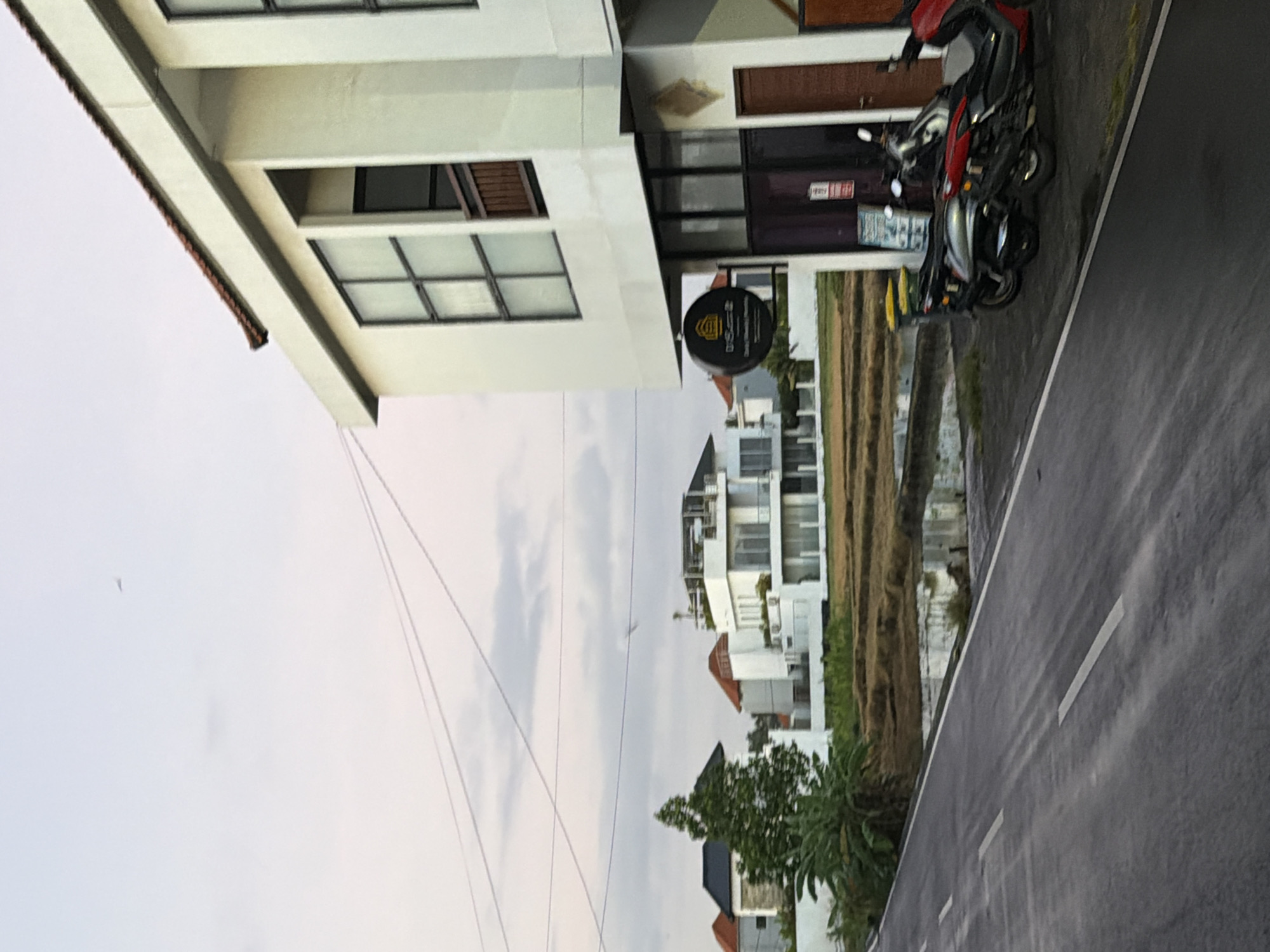 The image depicts a street scene in Mengwi, Indonesia. A two-story building with white exterior walls and dark-framed windows is visible on the left. A motorcycle is parked near the building's entrance. A small sign hangs outside the building. Beyond the building and across a narrow strip of land is a cluster of white residential buildings. A paved road with white lane markings runs along the right side of the image. The sky is overcast. Power lines are visible across the sky.