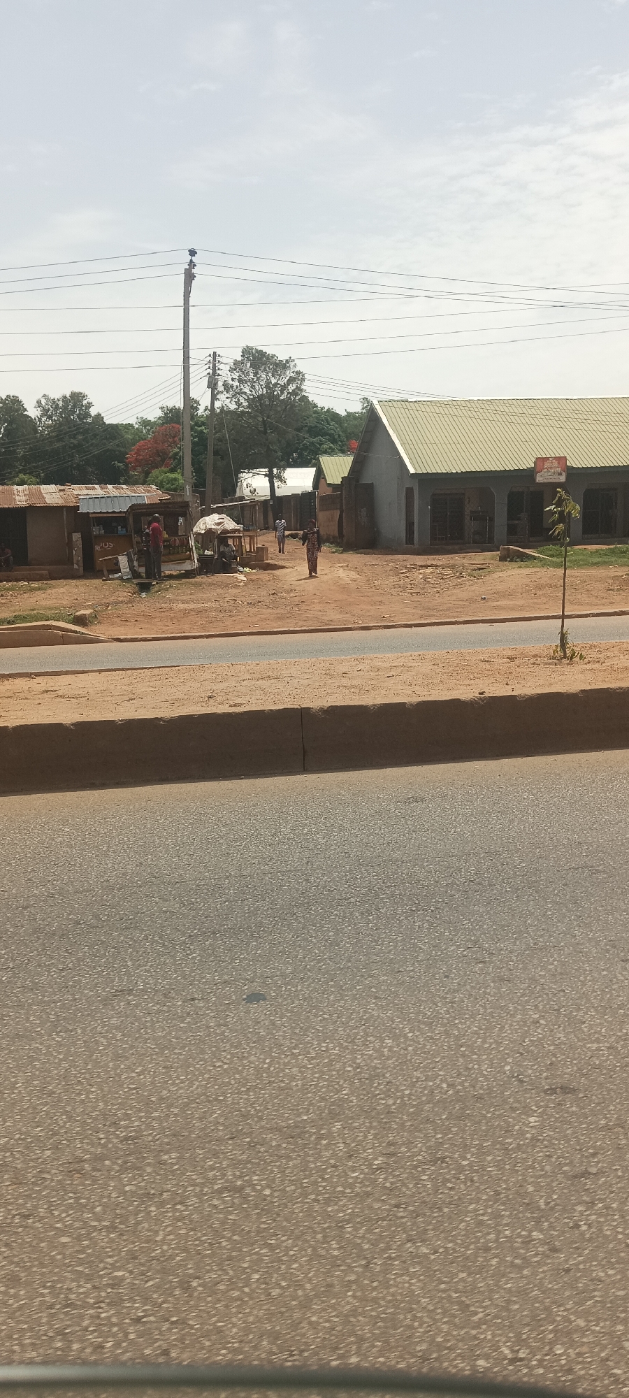 The image depicts a street scene in Bukuru, Nigeria, with the foreground dominated by an asphalt road and a raised curb. Across the street, several informal structures, possibly market stalls or small shops, line the dirt road. People are present, some standing near the structures and others walking. A utility pole with wires is visible. A building with a pitched roof is visible on the right. The sky is overcast.