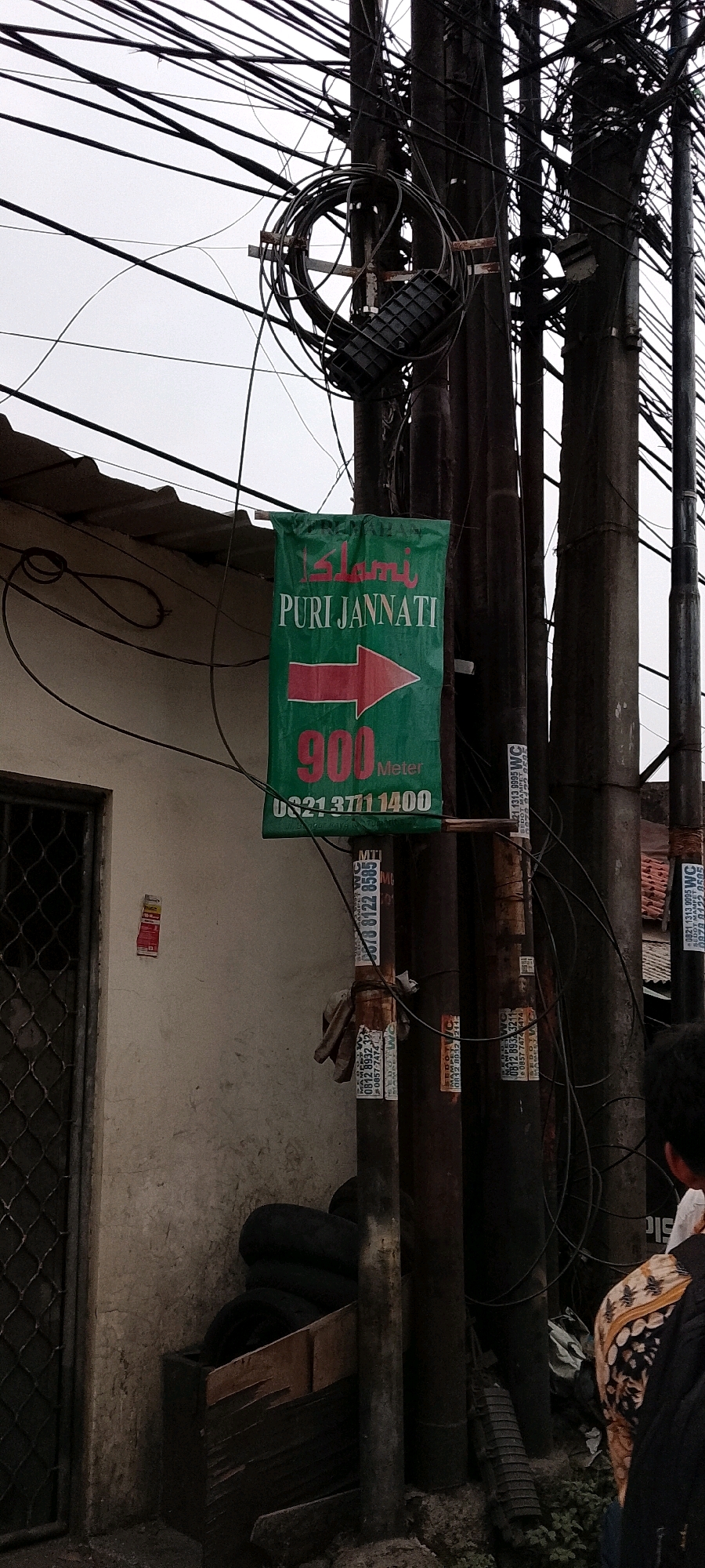 The image depicts an outdoor scene, likely in the city of Depok, Indonesia, characterized by a prominent utility pole adorned with a dense network of electrical and telecommunication wires. Attached to the pole is a green directional sign with red text indicating "Islami PURI JANNATI" and a red arrow pointing to the right. Below this, it displays "900 Meter" and a phone number "0821 3711 1400." Additional smaller stickers and tags are affixed to the utility pole. To the left of the pole, a building wall with a textured finish is visible, featuring a window with a metal grate and a section of a tiled roof. A wooden crate containing several tires is placed near the base of the pole. In the foreground, a person's back is visible, wearing a patterned shirt and a backpack. The overall environment appears somewhat cluttered with various urban infrastructure elements.