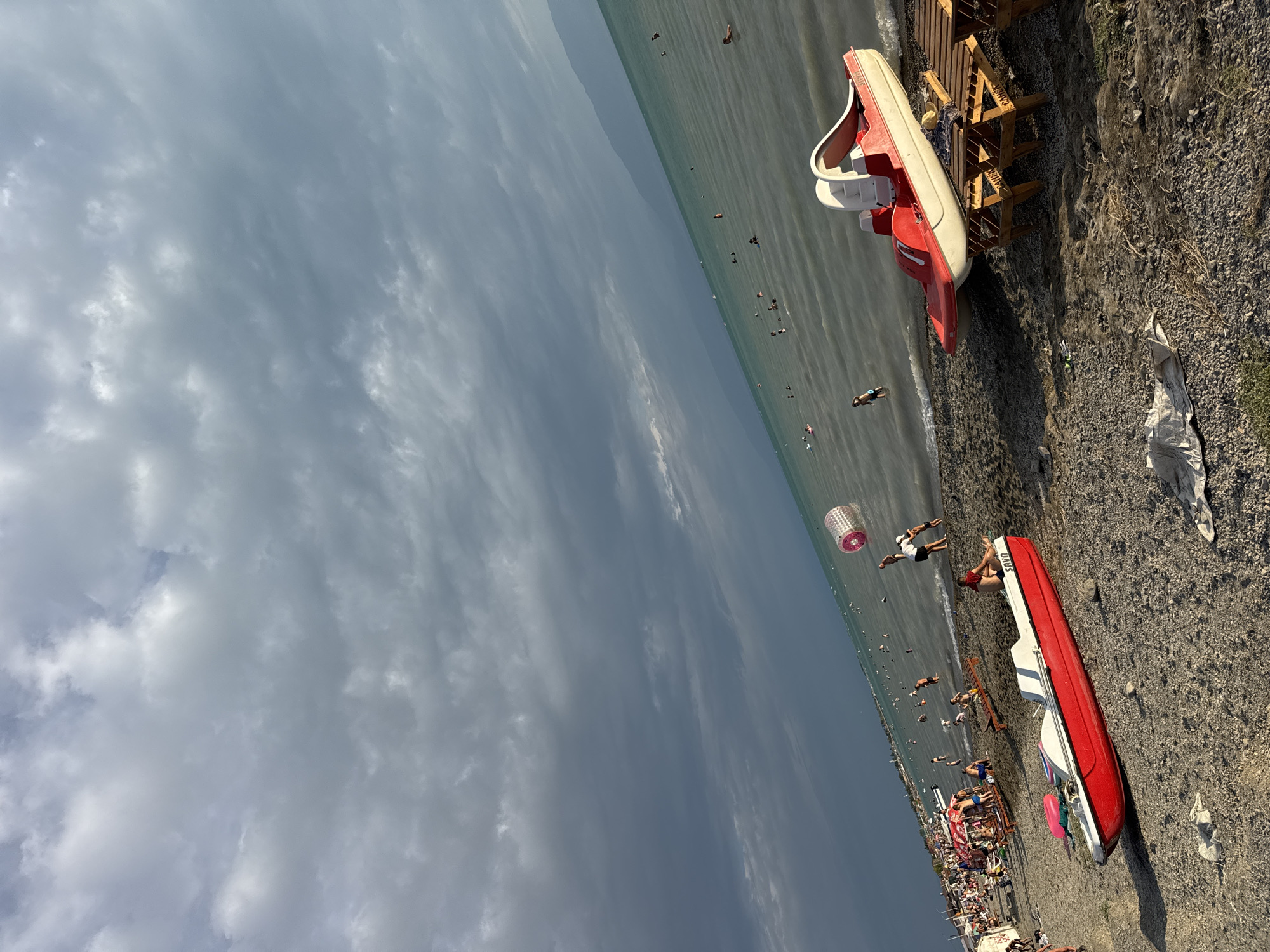 The image depicts a crowded beach scene with a shoreline, ocean, and cloudy sky. In the foreground, a red and white pedal boat is parked on the pebbly beach, with two wooden sun loungers nearby. Further back, numerous people are visible swimming and relaxing in the ocean. Along the beach, there are umbrellas and people gathered, suggesting a lively recreational area. The sea is a tranquil turquoise, meeting a distant mountain range under a dramatic sky filled with dark, heavy clouds. The overall impression is of a popular summer destination. The location is identified as Zharbulak, Kazakhstan.