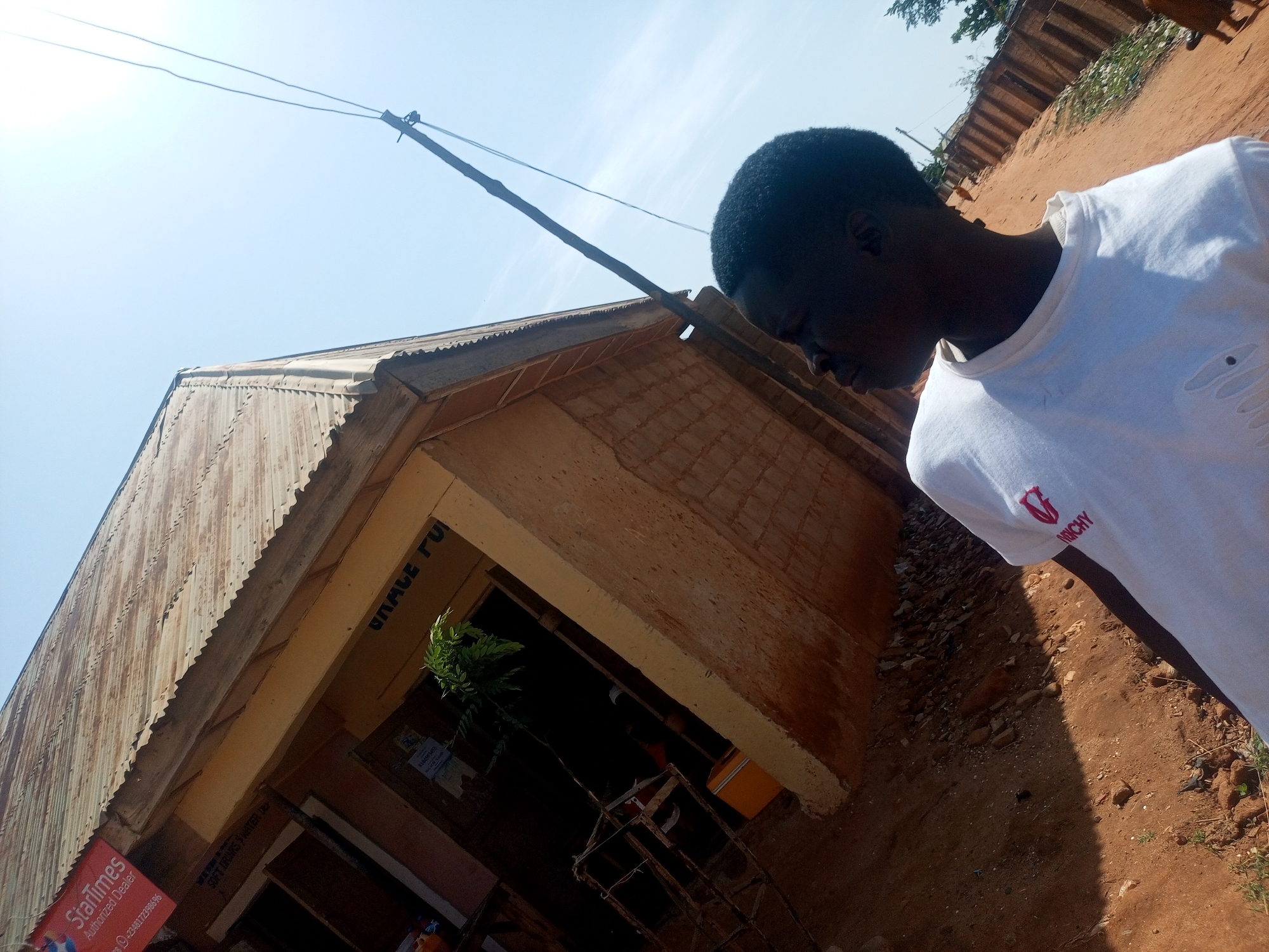 The image presents a sun-drenched outdoor scene in Tempure, Nigeria, captured with a Dutch tilt, looking up towards a building and the sky.
In the right foreground, a young person, likely male, is visible from the chest up. He has dark, short curly hair and dark skin, and is wearing a white, short-sleeved t-shirt with small distressed areas on the shoulder/sleeve. A red logo, possibly "GIVENCHY," is printed on the left chest. His head is angled downwards or away from the camera, obscuring his facial expression.
Behind the person is a single-story building constructed from light brown, possibly mud or plastered, material. It features a weathered, corrugated metal roof that shows signs of rust. Above the building's entryway, dark painted text partially reads "GRACE FU," suggesting a business name. Inside the dark entryway, a green leafy plant and other objects are discernible. A red sign near the entrance advertises "StarTimes Authorized Dealer" and includes a partial phone number starting with "23480." Another sign lists "SOFT DRINKS."
The ground is reddish-brown earth, characteristic of the region, with scattered rocks and sparse vegetation. A dirt path or road extends into the background. A utility pole with two wires crosses the upper part of the frame against a clear, bright blue sky, indicating bright daylight, likely midday or early afternoon. The scene depicts a commercial establishment in a semi-urban or rural setting.
