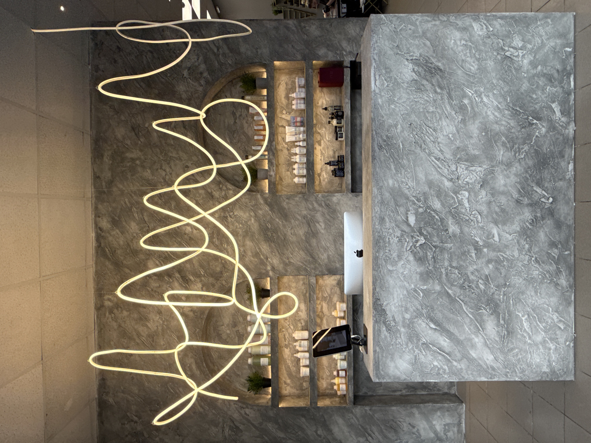 The image shows a high-angle view of an interior space in Votkinsk, Russia, focusing on a modern design aesthetic. The dominant feature is a large, irregularly shaped, abstract neon light fixture that spans across the left side of the frame. The walls are finished with a textured, gray concrete-like material. Built into the wall are several recessed shelves displaying various bottles and containers. There is a large, rectangular counter with a matching gray, textured surface that dominates the right side of the frame. On the counter, there's a white object (possibly a computer monitor) and a digital display. The lighting is bright, illuminating the textures and objects within the space.