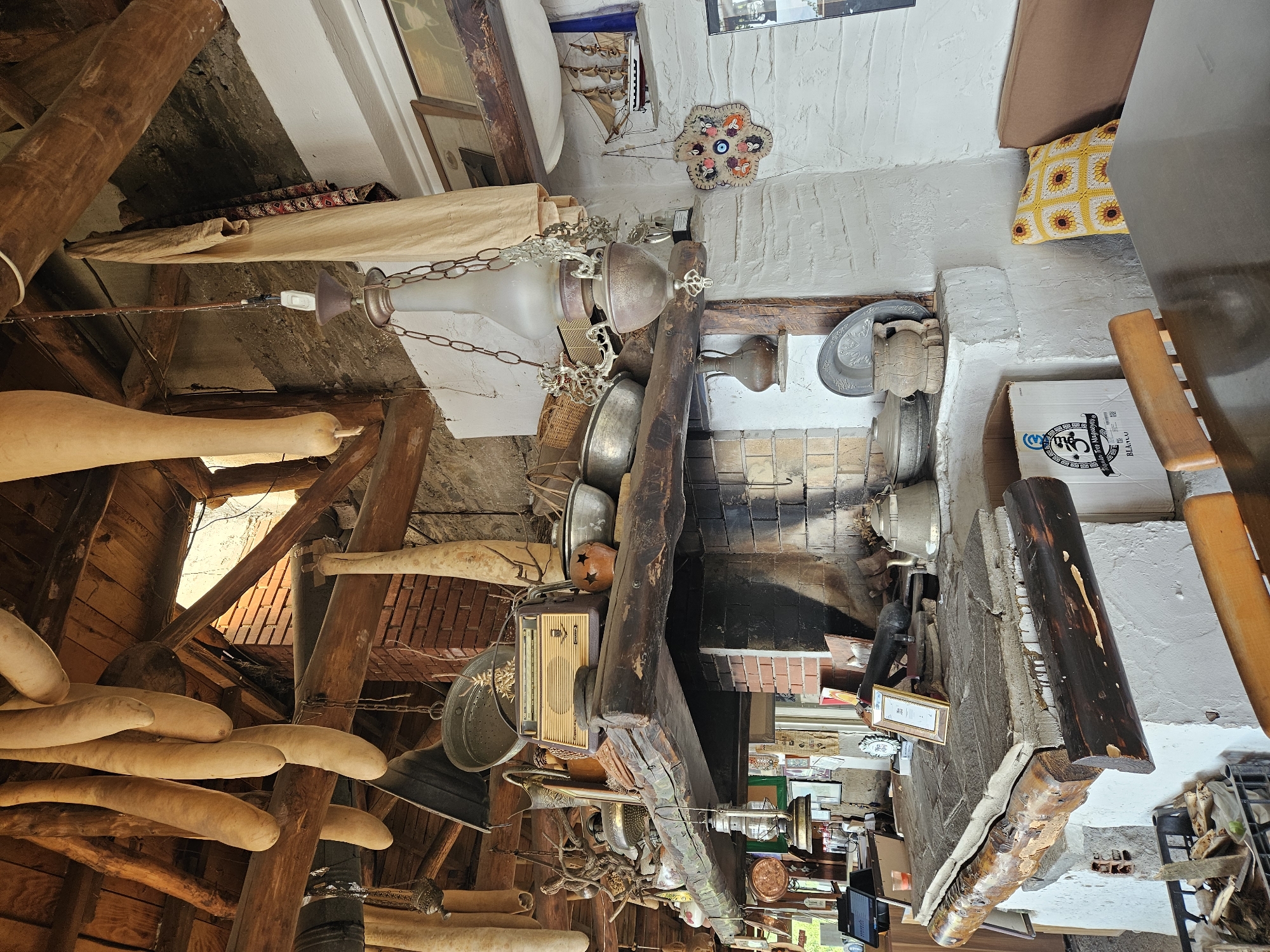 This image depicts the rustic interior of a establishment in Tire, Turkey, characterized by traditional architecture and an abundance of decorative and functional objects.
The scene is dominated by a large, brick fireplace structure with a prominent, dark-stained wooden mantelpiece and a lower cooking surface. The ceiling features exposed, rough-hewn wooden beams from which numerous elongated, light-colored, possibly dried gourds or plant husks, are suspended. A vintage-style chandelier with a frosted glass shade and chains hangs centrally, with a wall-mounted light switch visible nearby.
On the mantelpiece, a vintage radio with a light-colored grille and dark casing is prominently displayed, surrounded by various metal pots, a textured ceramic vessel, and decorative dried branches. Below the mantel, the fireplace opening shows sooted bricks, and to the left, a large, flat stone slab serves as a cooking surface or hearth. On this slab, a metal kettle, a framed certificate, and other miscellaneous items are arranged. A wooden log accent runs along the front of this lower structure.
The walls are predominantly white-washed, contrasting with exposed brick sections of the fireplace and the dark wood beams. On the right wall, a model wooden sailing ship is displayed on a small shelf, and below it, a circular, multi-colored decorative wall hanging featuring several "evil eye" motifs is suspended. The left side of the room shows a glimpse of a modern electronic register system.
In the foreground, a dark, reflective wooden table is visible, flanked by wooden chairs. A seating area with a plain brown cushion and a yellow crocheted cushion adorned with sunflower patterns is against the far right wall. A cardboard box labeled "Tequila Tres Magueyes BLANCO" rests on the floor near the table, and a black crate filled with wood scraps and possibly tools is also visible on the floor to the left. Sunlight enters the space from an unseen opening towards the upper left, highlighting the rustic textures and objects.