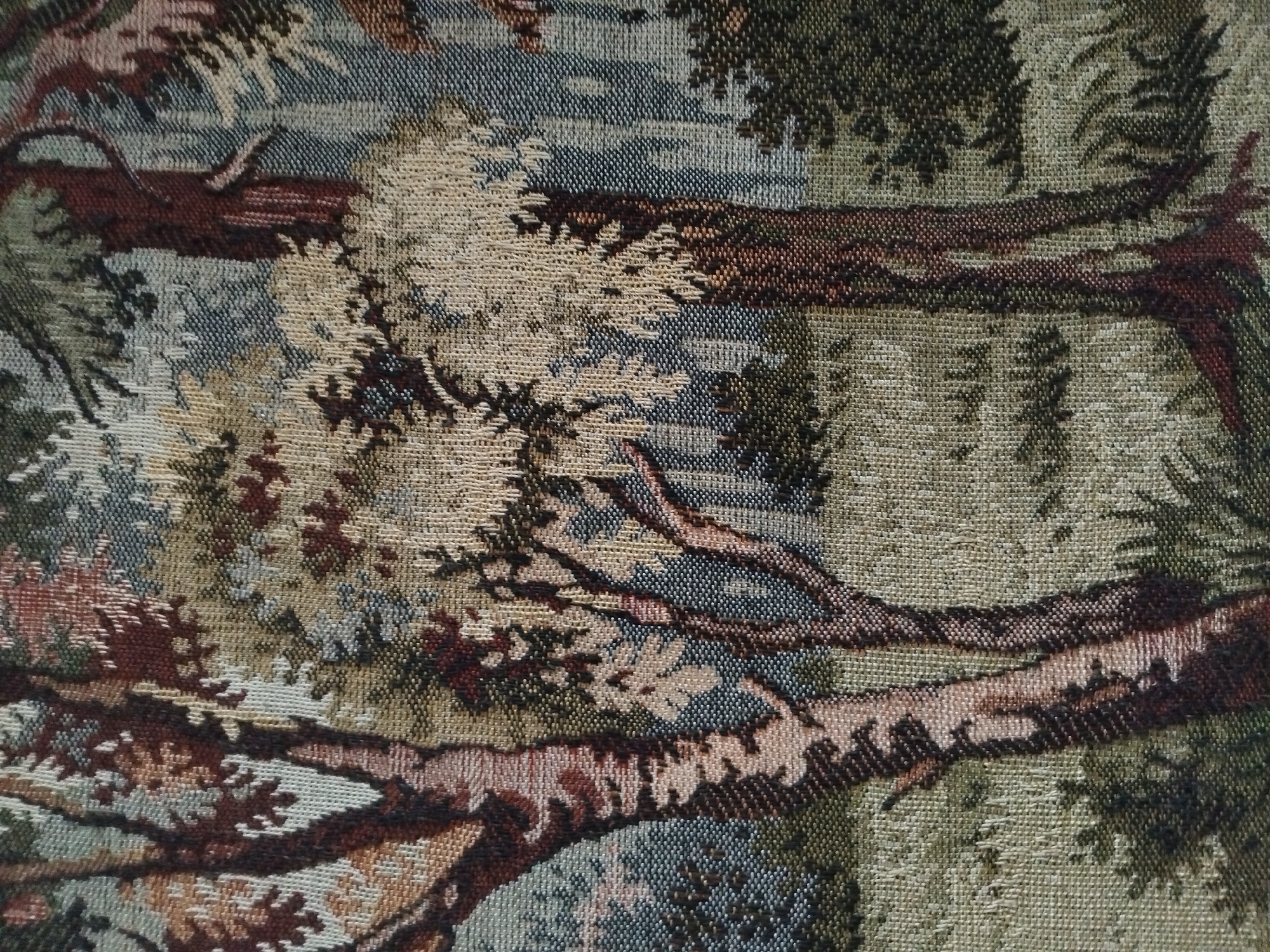 This image is a close-up, textured view of a tapestry depicting a forest scene. The tapestry is woven with threads of various earthy tones, including browns, greens, grays, and some muted reds. Dominating the foreground are the trunks and branches of trees, rendered with intricate detail. The texture suggests a woven fabric, possibly a wall hanging or upholstery. The background shows more trees and foliage, with a muted, hazy quality, creating a sense of depth. There are no people, animals, or discernible structures visible within the tapestry itself. The image does not provide any information about a specific location in Akhaltsikhe, Georgia, as it solely focuses on the artwork of the tapestry.