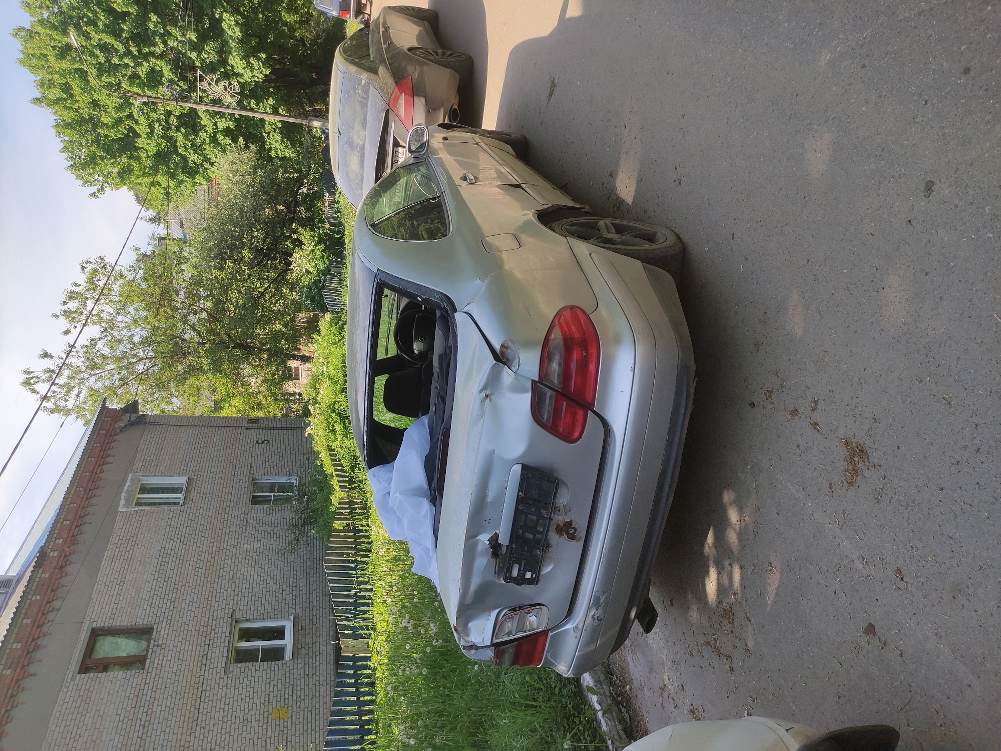 The image shows a damaged silver car parked on a street in Kratovo, Russia. The car's rear window is missing, and the trunk is heavily dented. A white cloth is visible inside the car. Another car is parked in front of the damaged car. A brick building and trees are in the background. The location appears to be a residential area.