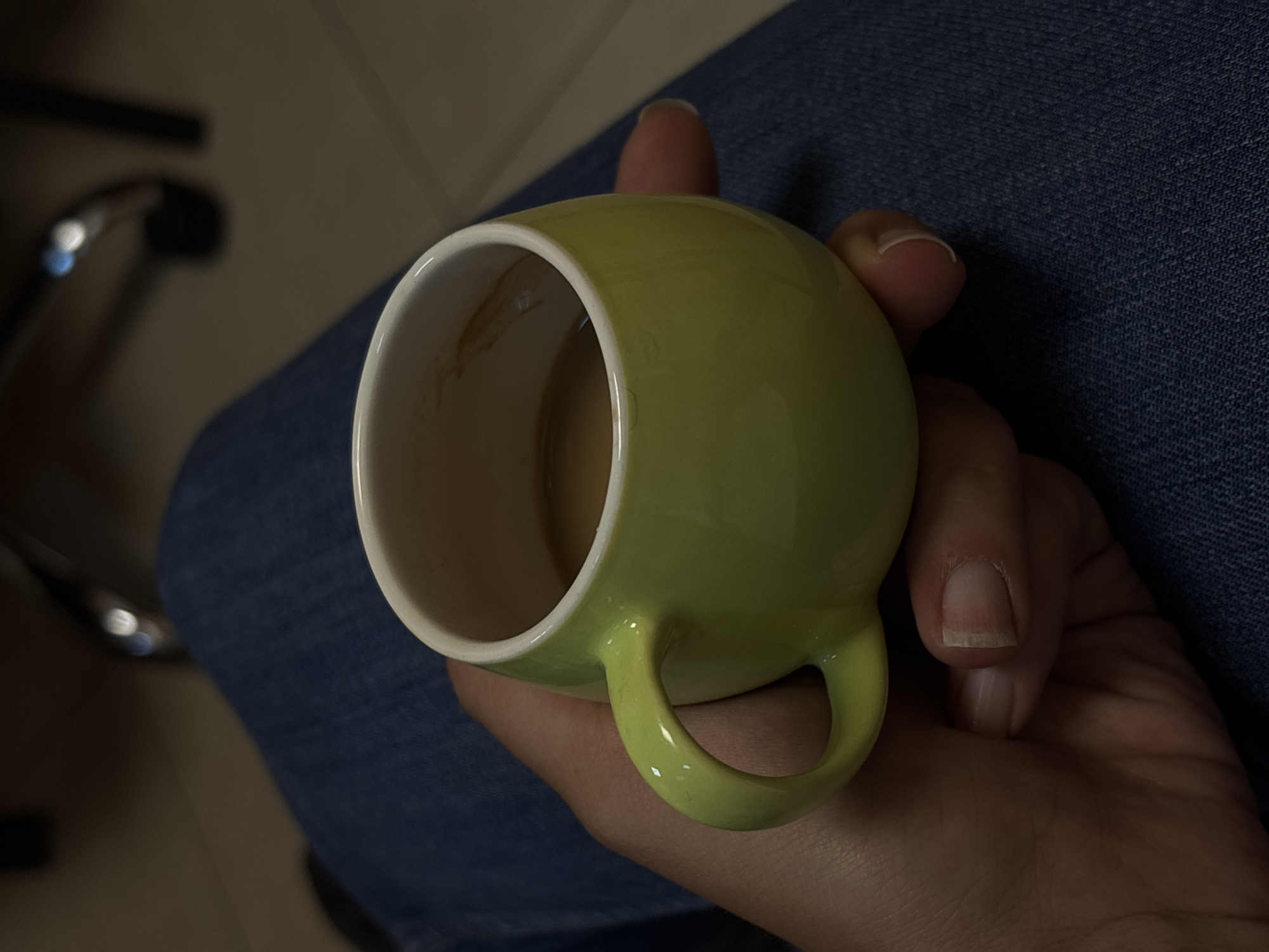 The image shows a close-up view of a hand holding a light green coffee mug filled with a light brown beverage, likely coffee or tea. The mug has a white interior with some staining from the drink. The hand is positioned to cradle the mug, with fingers wrapped around the handle and thumb resting on the rim of the mug. The person is wearing blue denim jeans, indicated by the texture and color of the fabric that fills the background. The lighting is dim, suggesting an indoor setting, possibly during the evening or in a dimly lit room. The scene is focused on the interaction between the hand and the mug, creating an intimate and personal feel. There is no visible text in the image. The overall impression is one of a quiet moment, perhaps a pause for a warm drink.