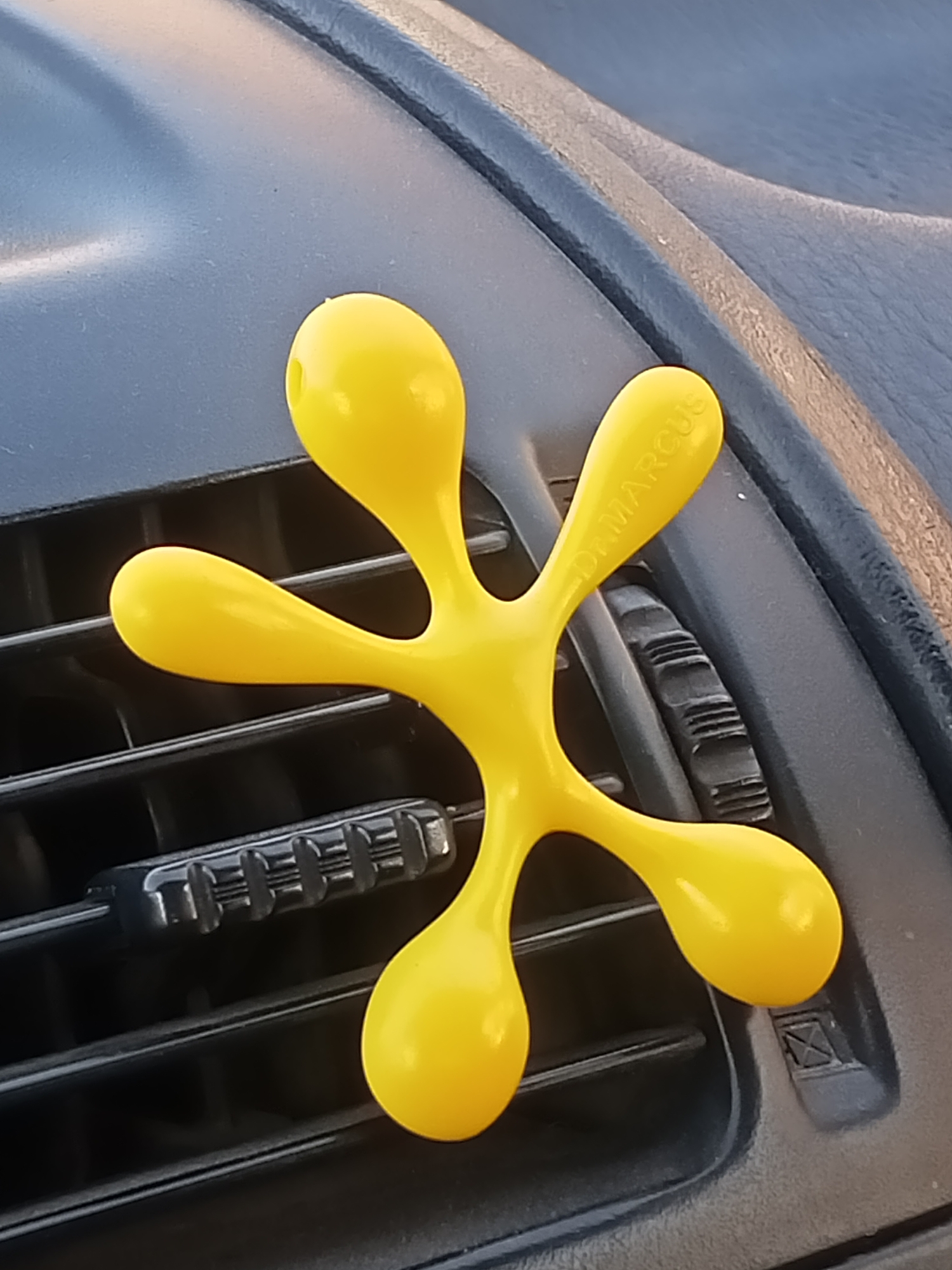 A yellow, anthropomorphic air freshener is attached to a black car vent. The air freshener has rounded limbs and a central body. The text "DR. MARCUS" is visible on one of its limbs. The car vent is made of black plastic with horizontal slats. The background is out of focus and appears to be the interior of a car. The lighting suggests it is daytime, and the scene is likely in Sokuluk, Kyrgyzstan, given the context provided.