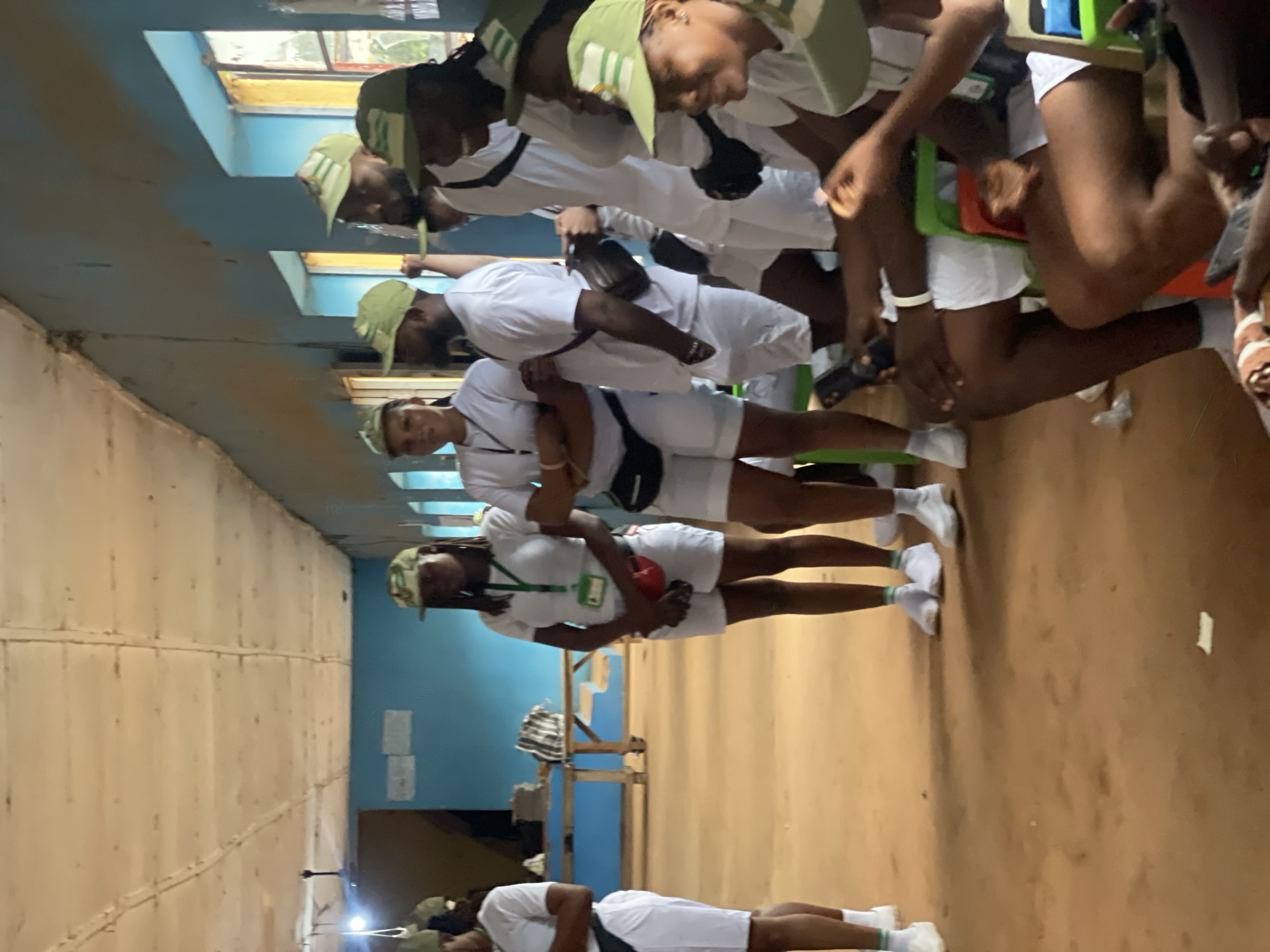 In Ise-Ekiti, Nigeria, an indoor scene captures a group of individuals in white uniforms and green caps, suggestive of a national service or youth program. The room features light blue walls, a light-colored, paneled ceiling, and natural light filtering through windows. The floor is a dusty, reddish-brown earth.
Several young adults are present, mostly standing, with some seated. They appear to be engaged in conversation or waiting. The individuals are dressed in white t-shirts, white shorts, and matching green baseball caps. Some wear lanyards with identification cards. One woman in the foreground is holding a red pouch around her waist. In the background, a wooden table with some items on it is visible. The overall atmosphere suggests a gathering or orientation session.