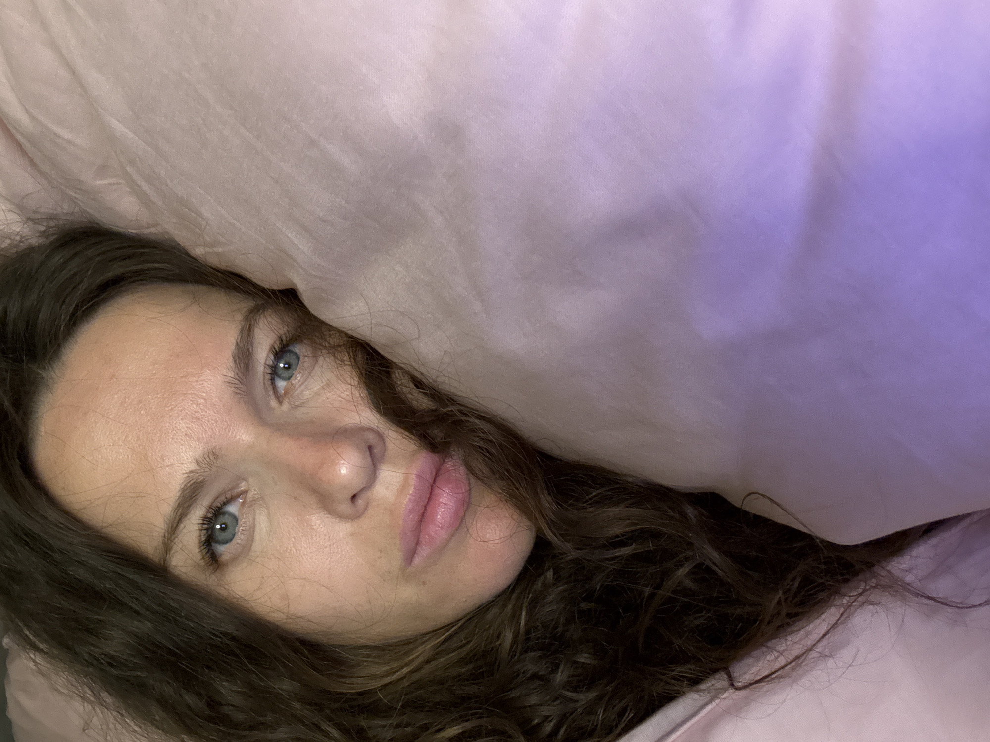 A close-up, overhead shot shows a woman lying in bed, resting her head on a pale pink pillow. Her blue eyes are open and looking at the camera. She has long, dark, curly hair that frames her face and cascades over the pillow. The woman's lips are slightly parted and appear to be a natural pink color. The background is dominated by the soft texture of the pillow, with a subtle gradient of purple light cast upon it. The setting appears to be a bedroom.