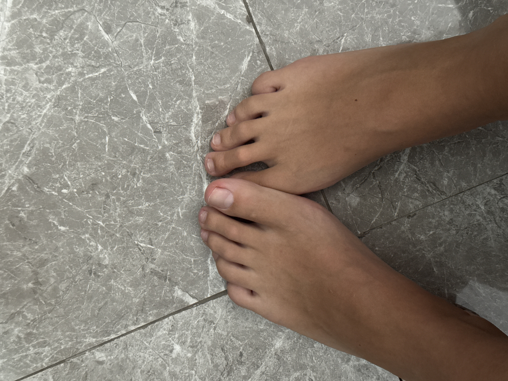 The image shows two bare feet on a grey tiled floor. The feet appear to be those of a young person. The toes are visible and well-formed. The floor tiles have a marbled pattern and are separated by grout lines. The background is out of focus. The image was taken indoors. There is no visual evidence to determine the location as North Kuta, Indonesia.