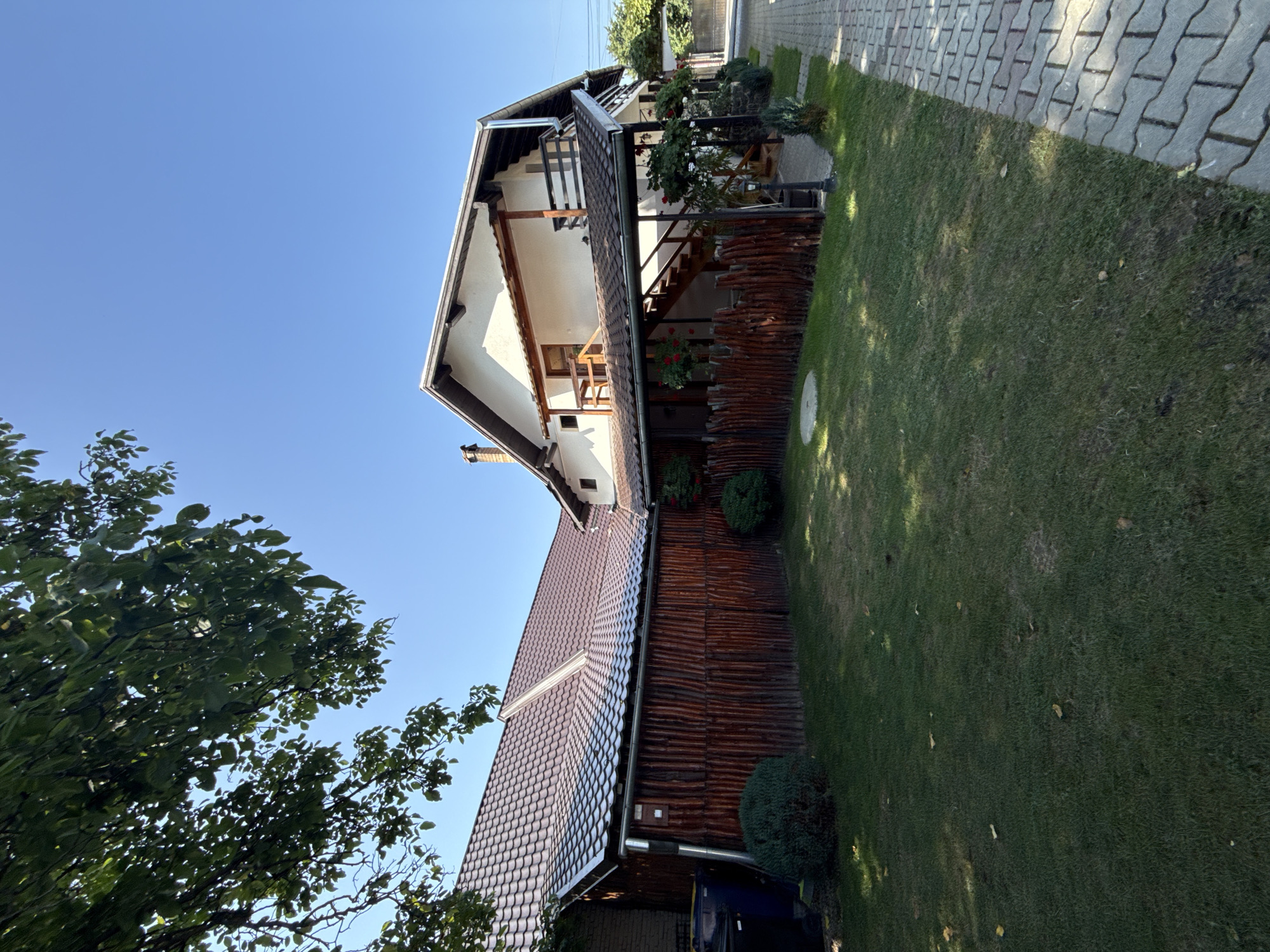 The image depicts a two-story house with a dark tiled roof, painted white on the upper level, and featuring wooden balconies. The lower part of the house is clad in vertical wooden planks, creating a rustic aesthetic. A wooden fence surrounds the property. To the left of the house, there are two large dark-colored trash bins. The yard is covered with green grass, with some scattered leaves. A paved walkway made of interlocking gray stones runs along the right side of the property. A large tree with green leaves partially shades the upper left portion of the image. The sky is clear and blue. The scene appears to be a residential area, possibly a guesthouse or a private home. The location is identified as Cârțișoara, Romania.