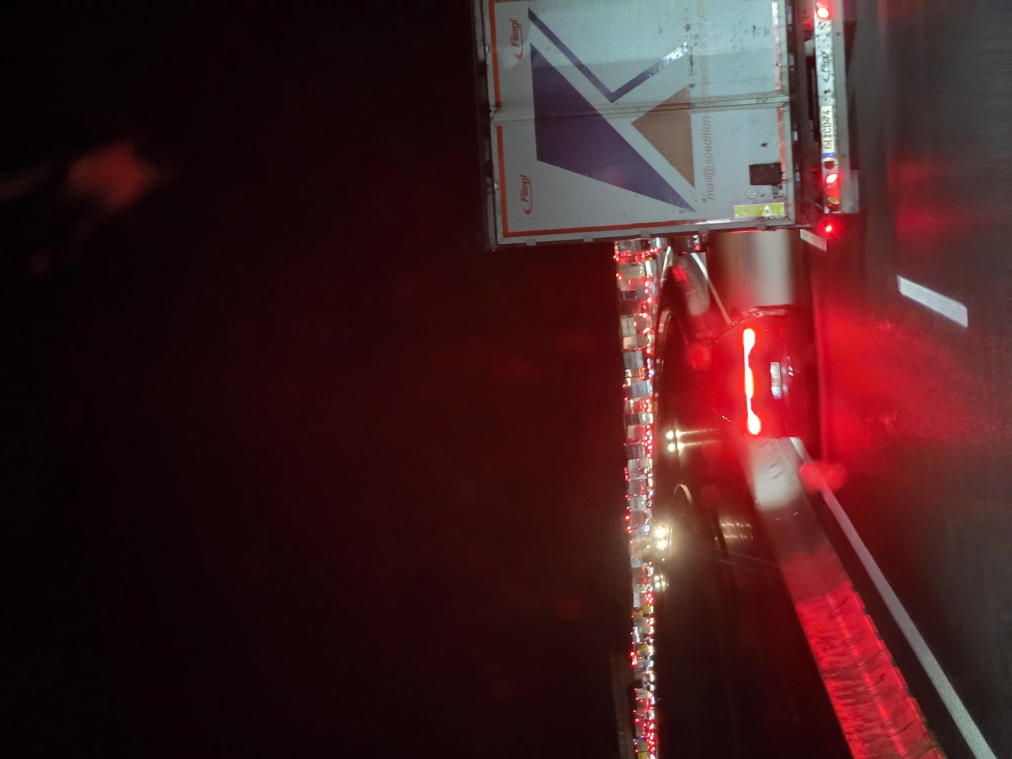 The image depicts a nighttime scene of heavy traffic congestion on a highway. In the foreground, a red car with its brake lights illuminated is stopped in traffic. To the right, the rear of a white truck with a large "K" graphic and the logo "Fliegl" is visible. The truck displays text that appears to be an email address: "mail@spedition-..." and a license plate that reads "BLCD-74". In the background, a long line of trucks and cars are stationary, with their red taillights creating a glowing effect against the dark, possibly wet, road surface. The sky is dark, indicating it is night. There is no indication of people's presence or specific interactions beyond the stopped vehicles. The location is identified as Uettingen, Germany, likely on a highway.