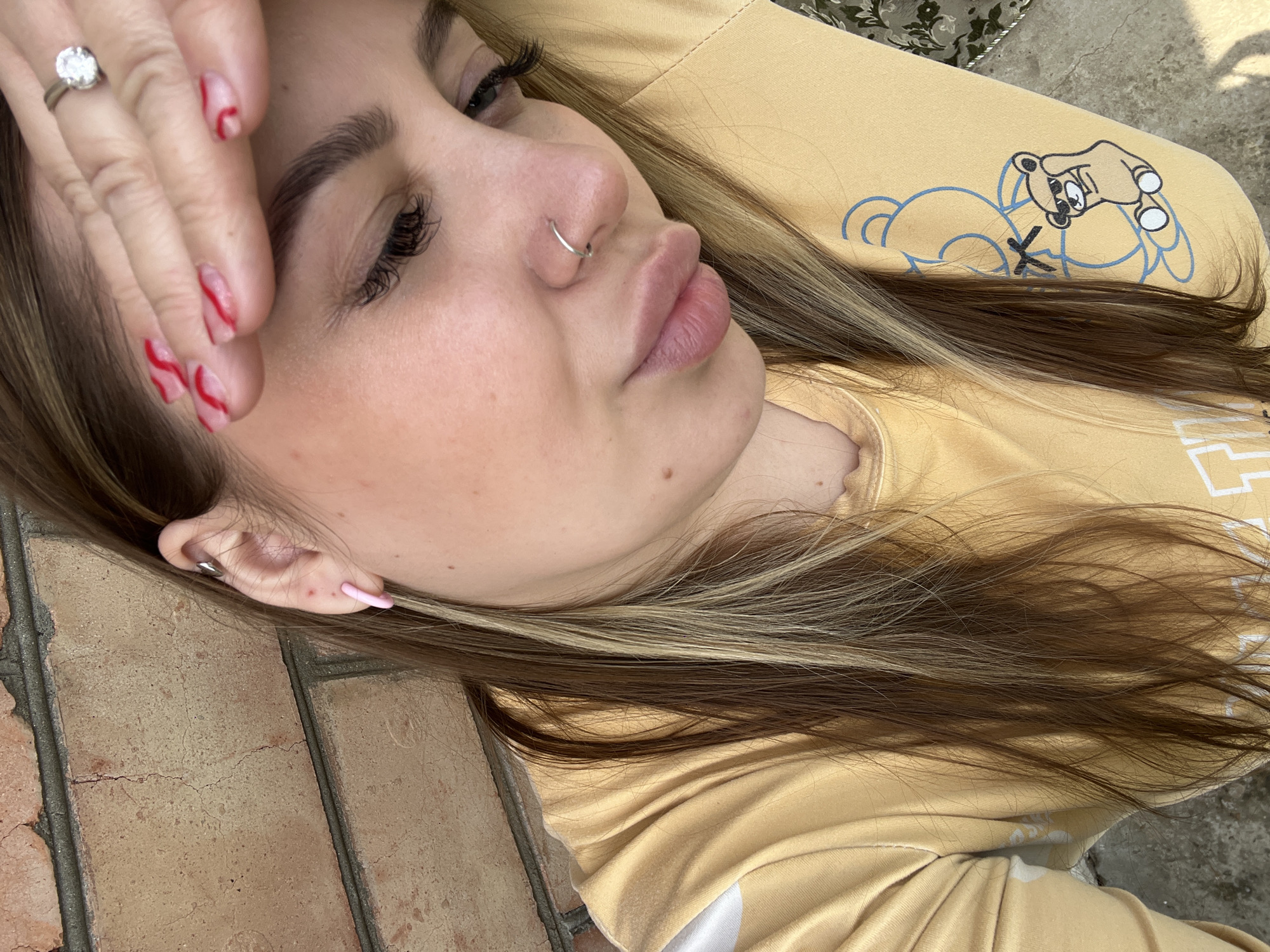 A close-up, slightly angled view of a woman's face with part of her body visible. Her hand, adorned with a diamond ring and red nail art, is held near her forehead. She has a nose ring, styled eyebrows, and long eyelashes. Her hair is highlighted, with a mix of blonde and brown strands. She's wearing a yellow sweatshirt, which has a cartoonish graphic on the sleeve. The background consists of brick flooring and another type of stone flooring. There is no information to confirm this image was taken in Novopokrovskaya, Russia.