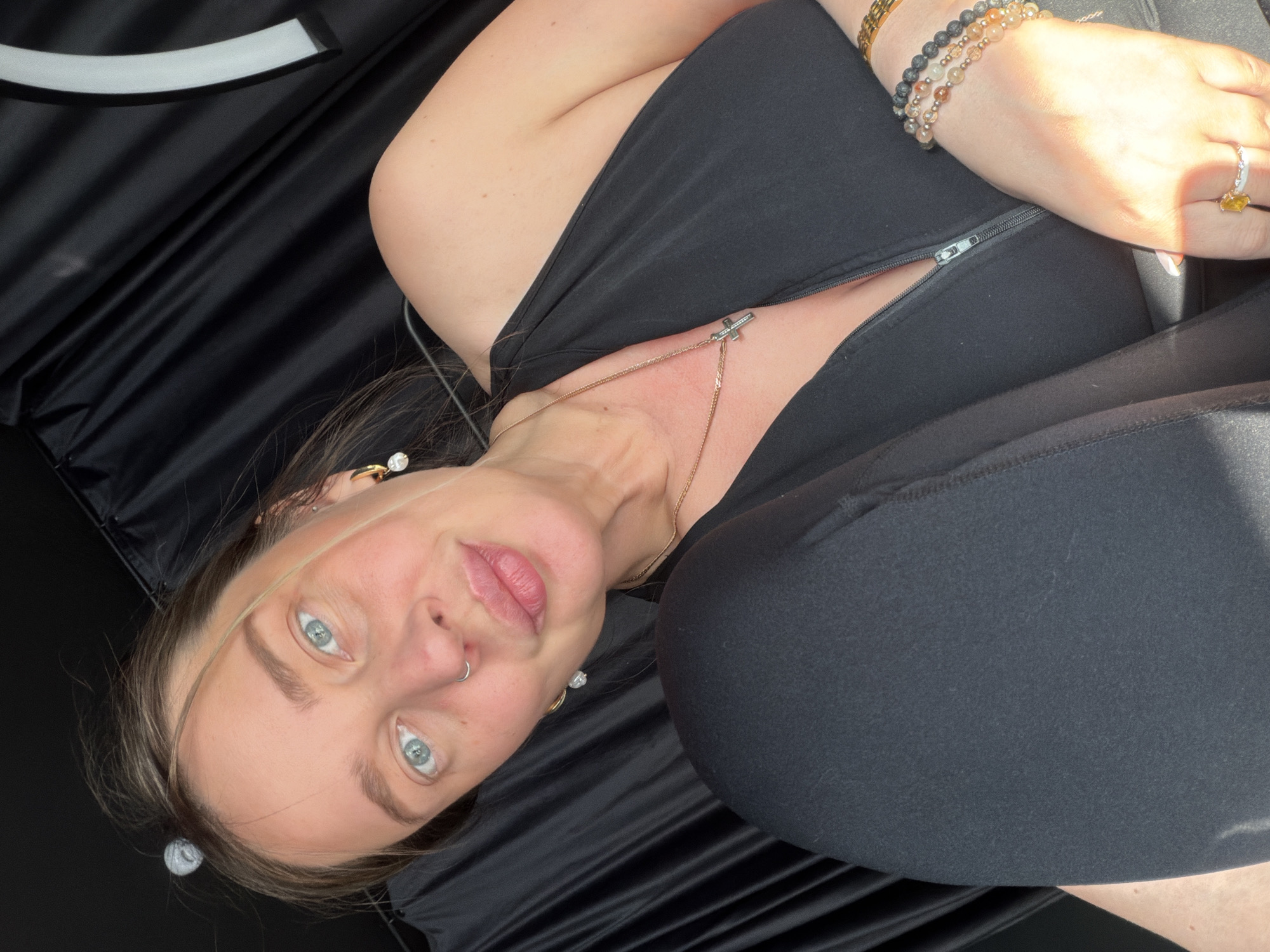 A woman with light blue eyes and light brown hair is pictured laying down on her side, with her face towards the camera. She is wearing a black top, black leggings, and several bracelets on her wrist. She is also wearing jewelry on her neck, ears, and nose. The background appears to be dark and flowing fabric, with a white curved object visible at the top left. The lighting suggests it is daytime, with sunlight illuminating the woman's arm and hand. The woman's expression is neutral, and her gaze is directed at the camera. There are no visible text or specific location cues in the image.