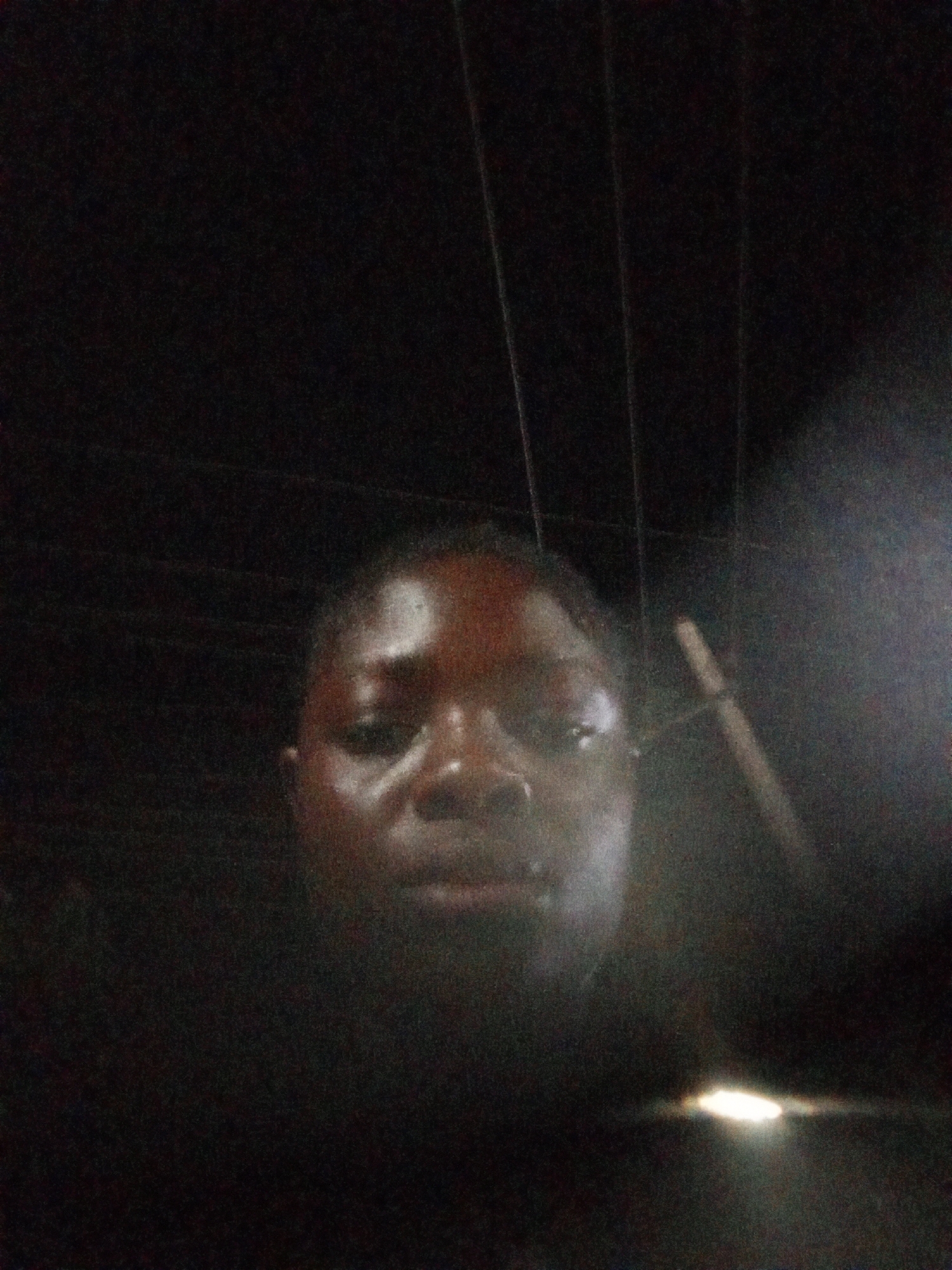 A dimly lit, blurry selfie of a person with dark skin. The individual is centered in the frame, and only their head and shoulders are visible. In the background, there are dark vertical lines, possibly utility poles or wires. A bright light source is visible in the lower right corner. The location, Mbribit, Nigeria, is not discernible from the visual information.