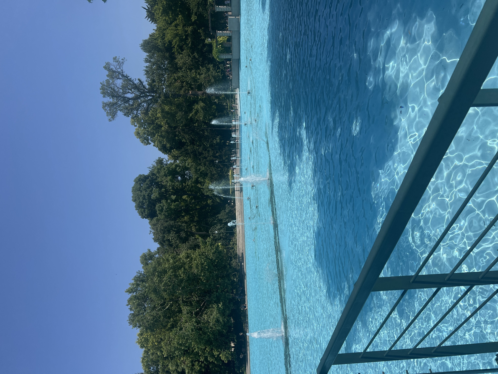 This is a full-frame, eye-level, outdoor shot of a large, bright blue public swimming pool or decorative fountain area in Plovdiv, Bulgaria. The water is calm with light reflecting off its surface, creating rippling patterns. Several small fountains are active within the pool, sending water jets upwards. In the background, lush green trees form a dense line, suggesting a park or garden setting. A dark metal railing is visible in the foreground, partially obscuring the view of the water. The sky above is a clear, vibrant blue. There are some people visible in the distance, sitting or walking along the edge of the pool area.