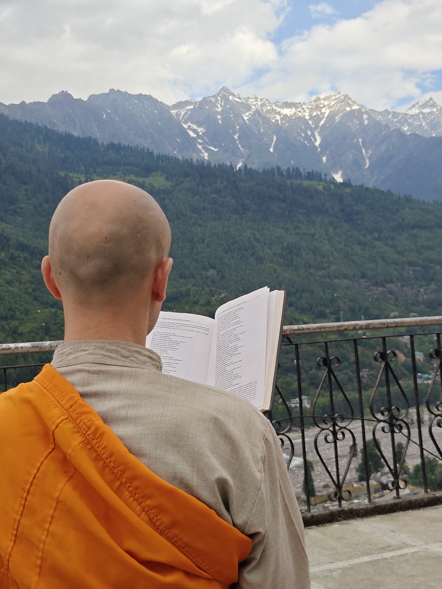 In Vashisht, India, a person with a shaved head, wearing a light-colored tunic and an orange shawl draped over their left shoulder, sits facing away from the camera. They are holding an open book and appear to be reading. The background features a balcony with a decorative metal railing. Beyond the balcony, a lush green mountain slope dotted with trees leads up to snow-capped mountains under a cloudy sky. The scene suggests a serene moment in a mountainous, natural environment.