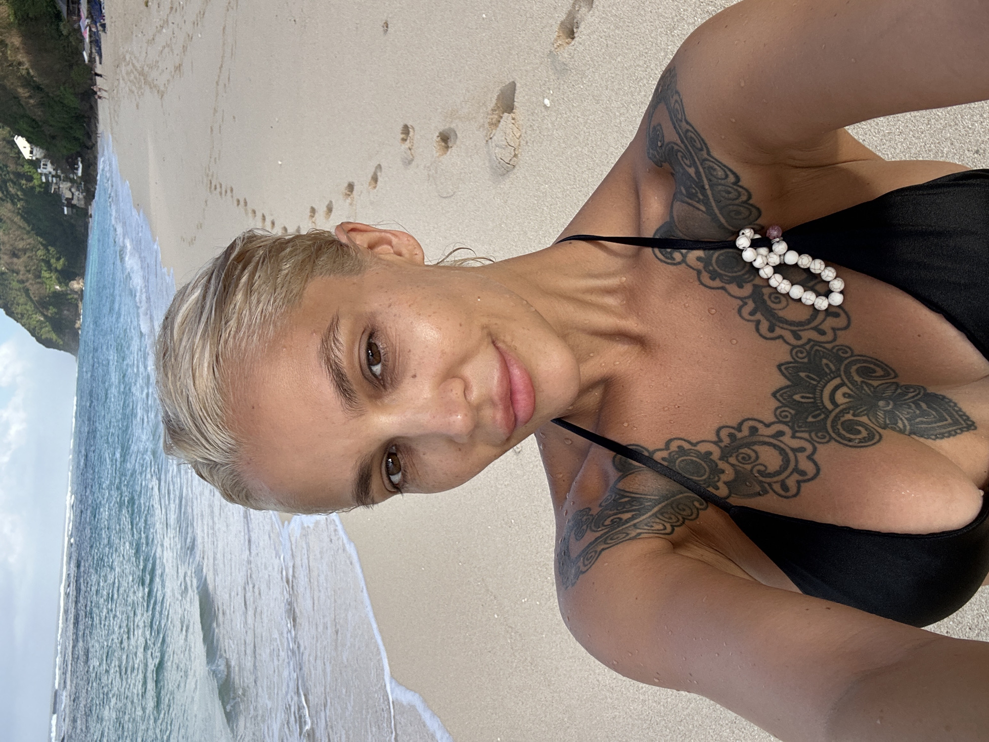 In Kuta Selatan, Indonesia, a blonde woman in a black bikini stands on a sandy beach. The ocean waves gently lap the shore behind her. Her skin is wet, and she has tattoos on her chest and shoulders. She is looking at the camera with a slight smile. Footprints are visible in the sand behind her. In the distance, lush green hills rise from the coastline, with buildings dotting the landscape. The sky is overcast with some visible clouds.