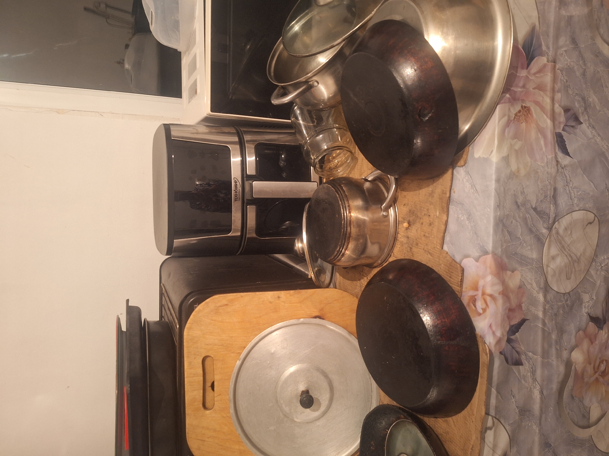 The image displays a collection of kitchenware, primarily pots and pans, arranged on a wooden surface. A coffee maker with the brand name "ТЕХНОМИР" is visible on the left. In the background, a microwave oven and a patterned tablecloth with floral designs can be seen. The setting appears to be a kitchen in a new building ("новостройка") in Kyrgyzstan. The lighting suggests it might be indoors, possibly in the evening. There are no people present in the image, and the overall mood is neutral, focusing on the objects.