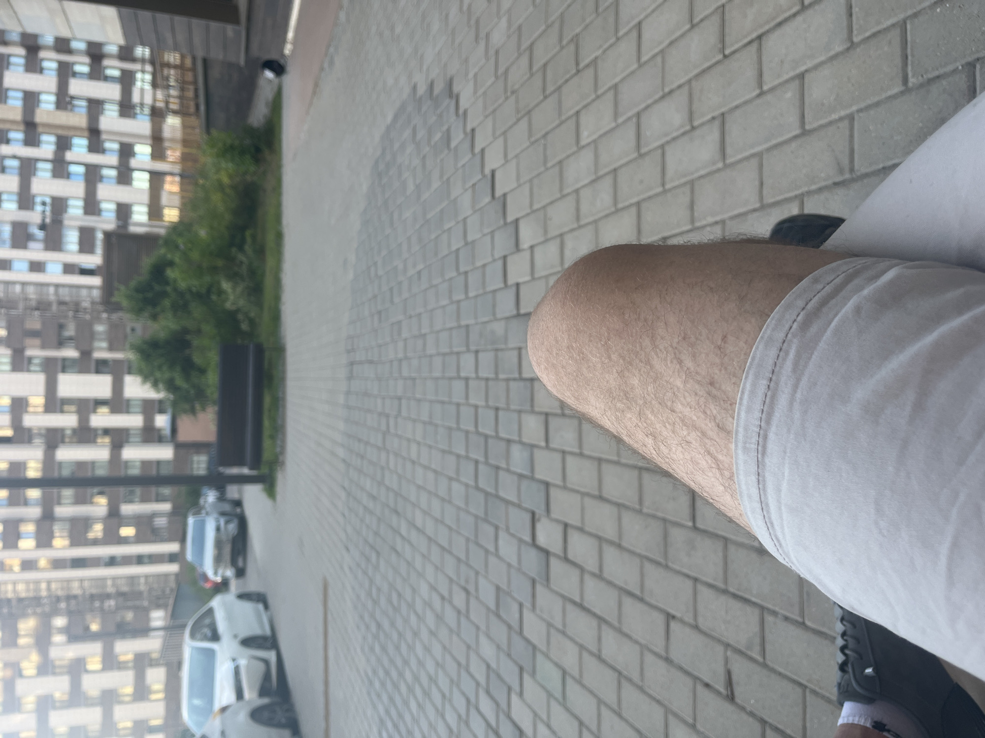 The scene is captured from a low, first-person perspective, looking forward from a seated position in a contemporary urban residential area.
In the immediate foreground, the upper right leg of an individual is prominent, showing visible leg hair and clad in light-colored shorts, likely beige or light grey. A white sock and a dark-grey lace-up shoe, belonging to the left foot, are partially visible in the lower-left corner.
The ground extending into the midground is a pedestrian surface paved with grey rectangular interlocking pavers arranged in a diagonal pattern. To the left, a paved road or driveway accommodates two white passenger vehicles. The closer vehicle is a white sedan, with its front-left quarter visible. Further back, a white SUV is observed, positioned head-on. On the right side of the paved area, a dark-colored park bench with a slatted design is present. Behind the road and the bench, a segment of green vegetation, consisting of trees and bushes, is visible.
The background is dominated by multiple large, multi-story residential buildings. These structures display a modern architectural style characterized by patterned facades that integrate light-colored (white or beige) vertical and horizontal panels with darker (grey or brown) sections and numerous windows. The lighting is consistent with diffused daylight. The environment is typical of a modern residential development found in cities like Khimki, Russia. No specific interactions or actions are discernible beyond the implicit observation by the individual whose leg is in the foreground.