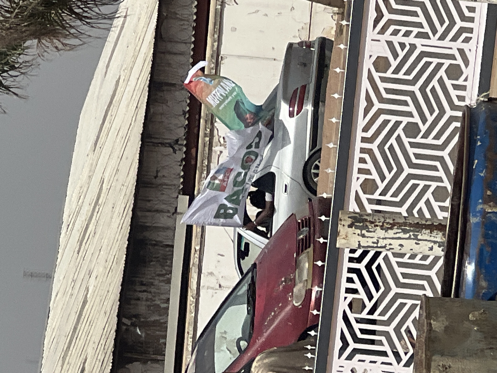 The image shows a white car with political banners hanging out of its windows. The banners are green, white, and red. One banner has the word "MUTUMWAN" printed on it. The car is parked next to a building with a white lattice design. The image was taken in Jos, Nigeria. There is a red car door near the white car. The background shows a cloudy sky.