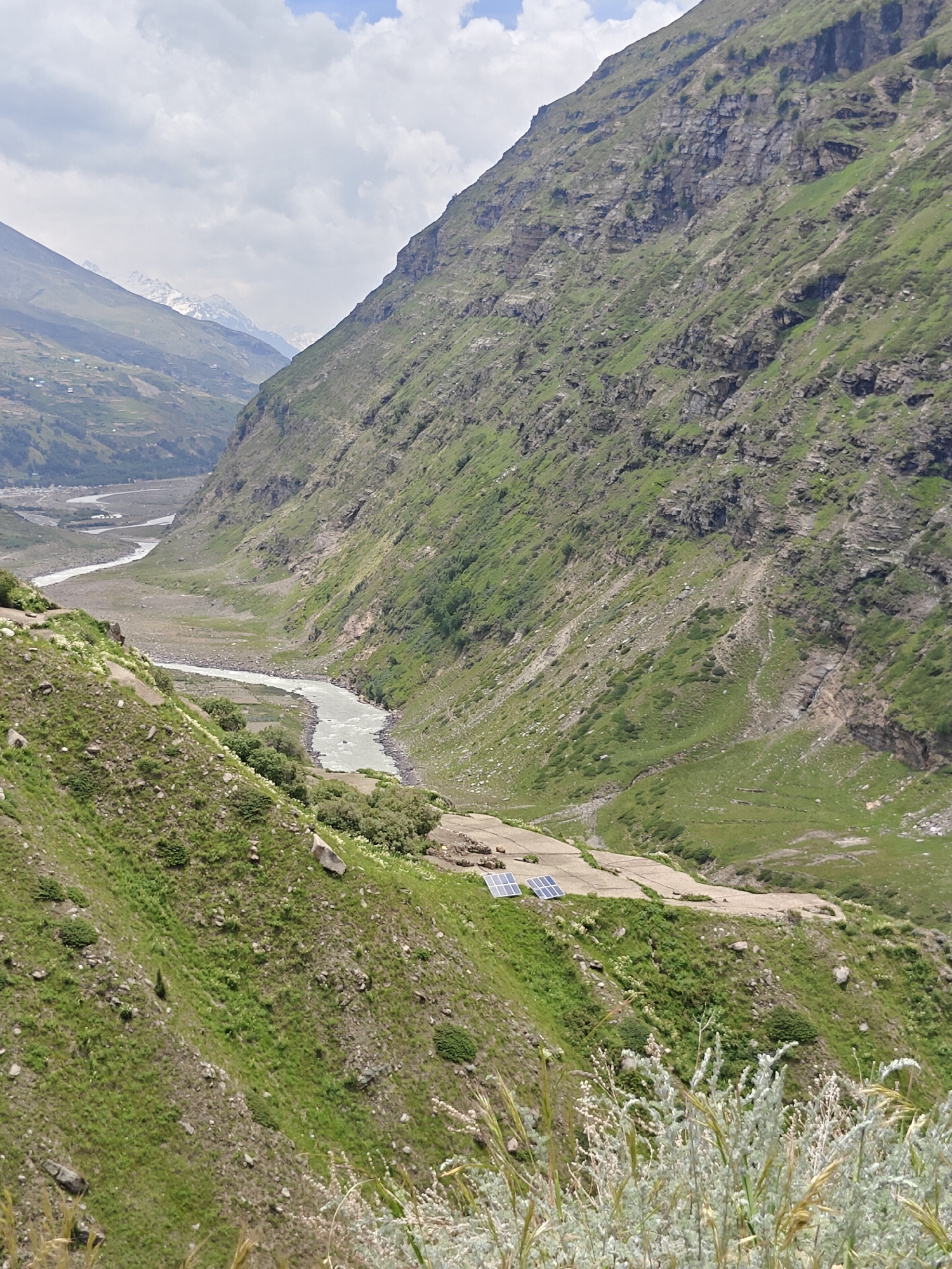 A wide-angle, high-angle shot captures a natural landscape in a mountainous region. The scene is dominated by steep, verdant slopes on either side of a winding river that flows through a valley. The river appears milky or silty. On the right side of the frame, a sheer rock face is visible, interspersed with patches of greenery and scree. The left side of the valley features gentler slopes, with evidence of terraced fields and some scattered structures at a distance. In the foreground, on a slightly elevated plateau of the left slope, a small installation of solar panels is present. The sky above is partly cloudy with a mix of white clouds and blue sky. The overall impression is one of rugged, natural beauty, with a hint of human presence through the solar panels and distant settlements.