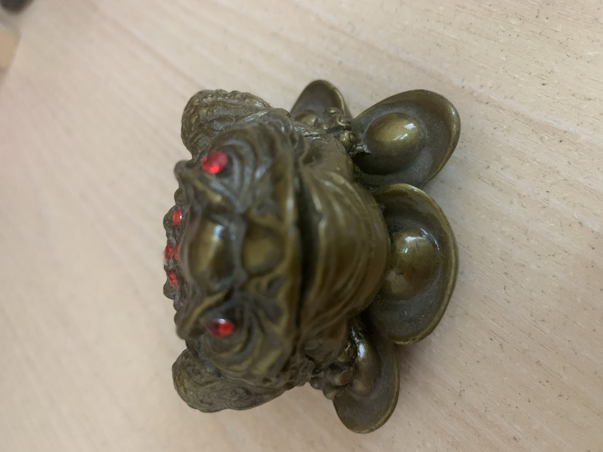 A bronze-colored Feng Shui Money Frog statuette with red eyes rests on a light-colored wooden surface. The frog appears to be sitting on a bed of Chinese ingots (yuan bao). The frog's head is textured, and the ingots have a smooth, rounded appearance. The image does not provide enough information to determine its location in Chaikovsky, Russia, or to identify any people, actions, or interactions. The environment is simply the wooden surface upon which the statuette sits.