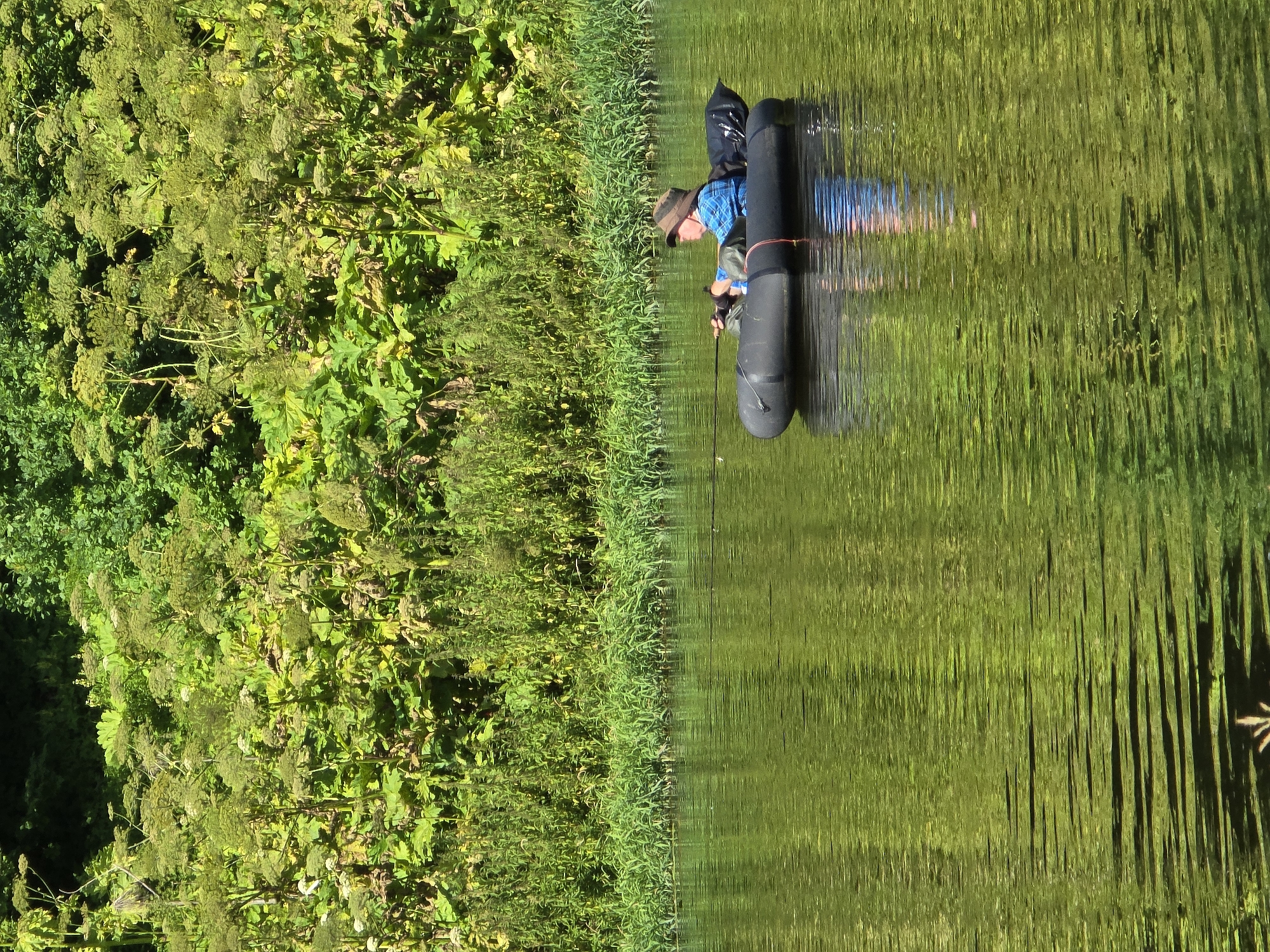 A person wearing a hat and a blue and white checkered shirt is fishing from a black inflatable boat on a body of water. The person is holding a fishing rod. Behind the boat, the bank of the river is covered in lush green vegetation. The water reflects the green of the foliage and the blue of the sky. The scene is lit by sunlight, casting reflections on the water. The image does not contain any specific landmarks or identifiable features that would allow for pinpointing a location within the city of Voinskaya Chast' Mchs, Russia.