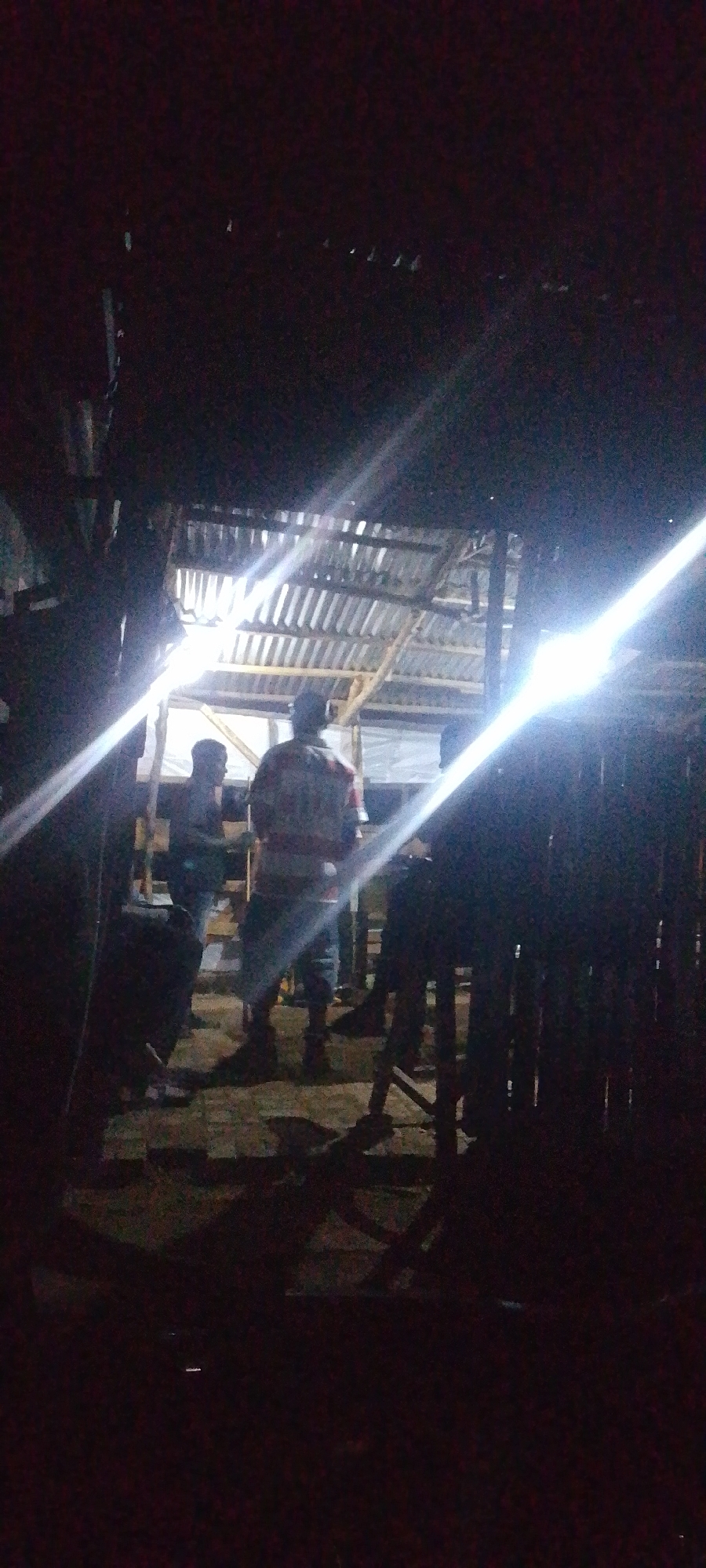 The image shows a dimly lit scene, possibly an outdoor gathering or construction site at night in Gura Top, Nigeria. Several individuals are present, silhouetted against bright artificial lights that create lens flares and streaks across the frame. In the foreground, a person wearing a patterned shirt and shorts is visible from the back, standing near a wooden fence. To their left, another person in a lighter colored shirt and dark pants stands partially obscured by the darkness. In the background, a corrugated metal roof supported by wooden beams suggests a makeshift structure. The overall impression is one of a casual, possibly impromptu, nighttime event or work in progress. The environment appears to be rustic and underdeveloped, typical of some areas in Gura Top.