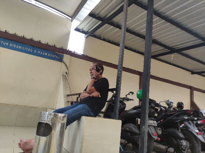 This image depicts a candid daytime scene in a semi-outdoor, sheltered area, likely in Kepanjen, Indonesia. The main subject is an adult man, appearing middle-aged, who is seated on a low, light-colored concrete ledge or platform. He is dressed in a black t-shirt and blue jeans, with his arms crossed, and a watch visible on his left wrist. He is actively engaged in a phone call, holding a black smartphone to his right ear. His bare feet are visible, resting near a tall, metallic ashtray or waste bin positioned in front of him.

Behind the man, a cream-colored wall features a blue sign with white text. Part of the text is visible, reading "TUK DISABILITAS & KAUM RENTAN," which likely translates from Indonesian as "FOR PEOPLE WITH DISABILITIES & VULNERABLE GROUPS," indicating a dedicated or accessible space. Above the sign, along a brown ledge, anti-bird spikes are visible. A broom or mop handle leans against the wall to the left of the man.

To the right side of the frame, several motorcycles are parked, suggesting this area might serve as a waiting spot or a parking section. One motorcycle clearly displays a green helmet. The entire area is covered by a corrugated metal roof, supported by dark, sturdy pillars, providing shelter from the elements. The bright, diffused light from above indicates it is daytime. The overall setting is functional and utilitarian, reflecting a common urban or public space in Indonesia.