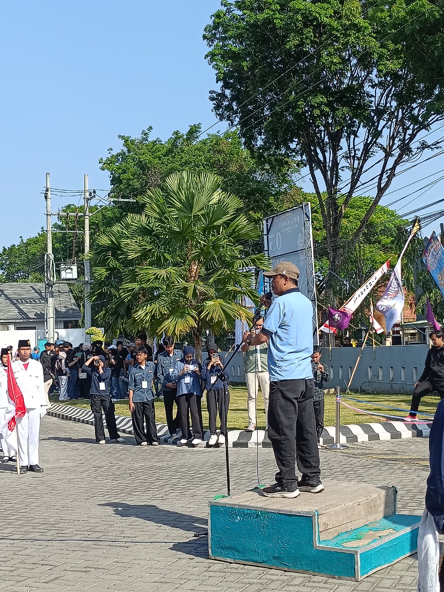 A man wearing a blue t-shirt, black pants, and a cap stands on a blue platform, speaking into a microphone. He is facing a crowd of people, some of whom are holding cameras and phones. The crowd is gathered on a paved area with a pattern of white and black stripes. In the background, there are trees, buildings, and flags. One flag, predominantly purple and white, displays the text "SMAN 5" (Senior High School 5). A white banner with Indonesian text is also visible behind the speaker. The scene suggests an outdoor event or ceremony taking place in a public space, possibly related to a school event in Kediri City, Indonesia. The sky is clear and blue, indicating a sunny day.