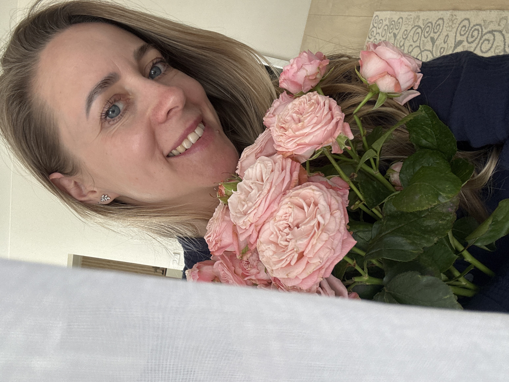 A woman with blonde hair and blue eyes smiles at the camera while holding a bouquet of pink roses. She is wearing what appears to be a dark blue top and a silver earring. The background includes a white surface in the foreground, a cream-colored wall and what may be a patterned rug on the right. There is no specific indication that the picture was taken in the city of Chaikovsky, Russia.