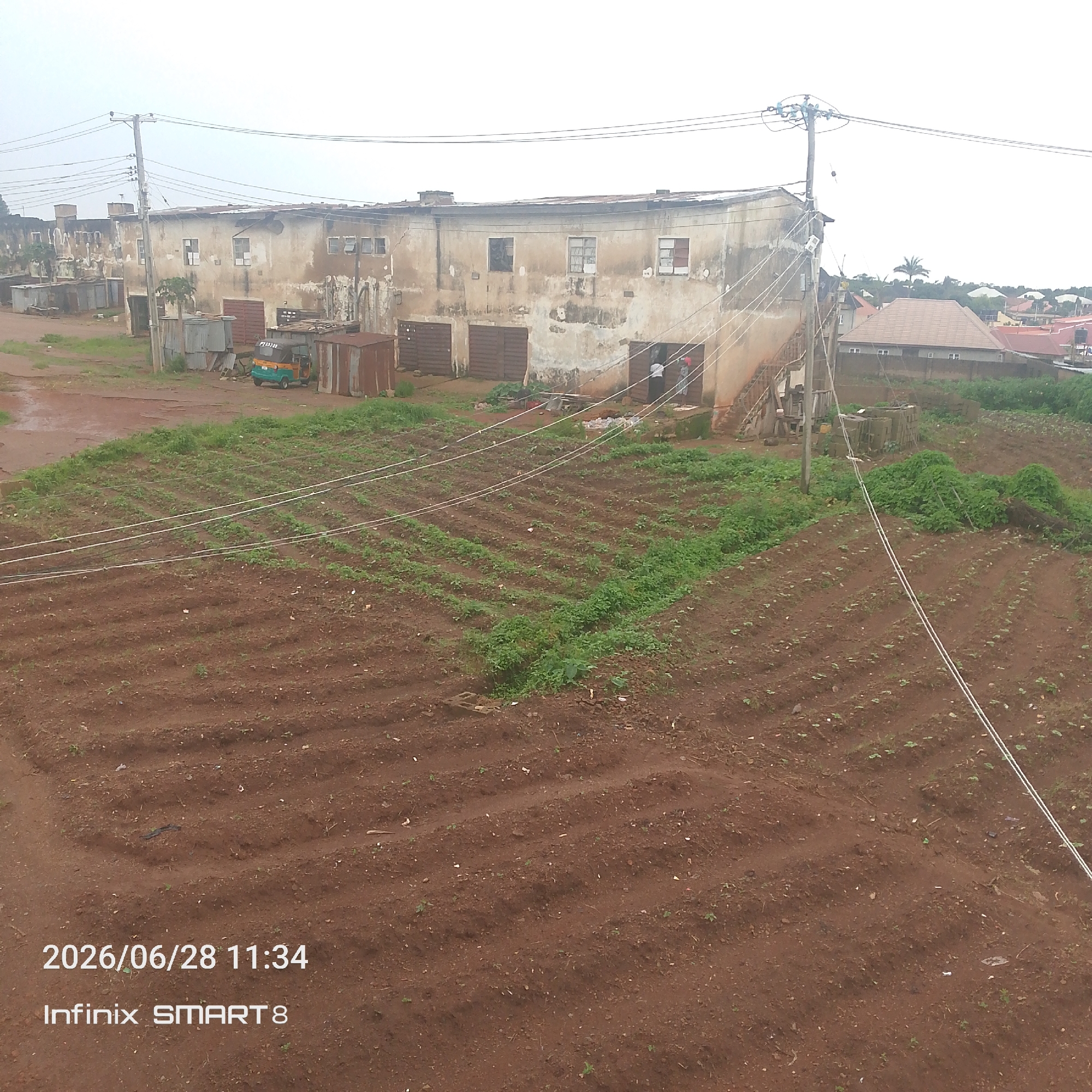 The image shows an aerial view of an urban area with residential buildings and agricultural plots. In the foreground, there are rows of crops planted in dark soil, with some green foliage visible. Several white wires run across the agricultural fields. In the background, a long, weathered building with multiple windows and doors stands. Adjacent to this building are smaller structures, some made of corrugated metal. A small, multi-colored auto-rickshaw is parked near these structures. Further in the background, residential houses with tiled roofs are visible, along with trees and vegetation, indicating a mixed urban and semi-rural landscape. The sky is overcast. A timestamp overlay at the bottom left reads "2026/06/28 11:34", and a phone model overlay reads "Infinix SMART 8".