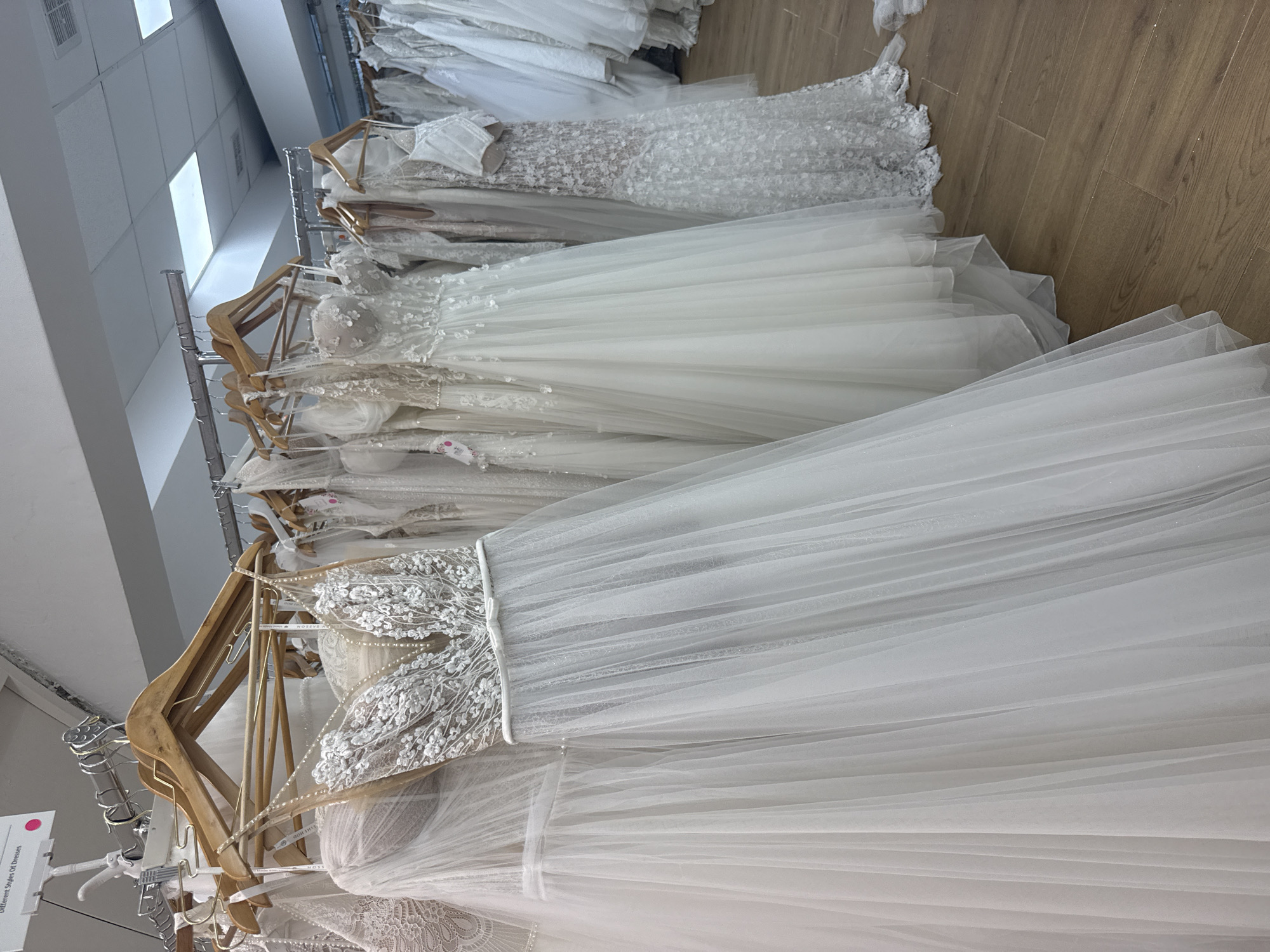 The image shows a row of wedding dresses hanging on a rack in what appears to be a bridal shop. The dresses are various shades of white and off-white, some with intricate lace or beaded detailing on the bodices. The skirts are voluminous and made of layered fabrics, likely tulle or chiffon. The dresses are hanging on wooden hangers, and the rack is located against a white wall with a wooden floor. The lighting is bright and artificial.
There is no information to suggest the location is Tel-Aviv, Israel.