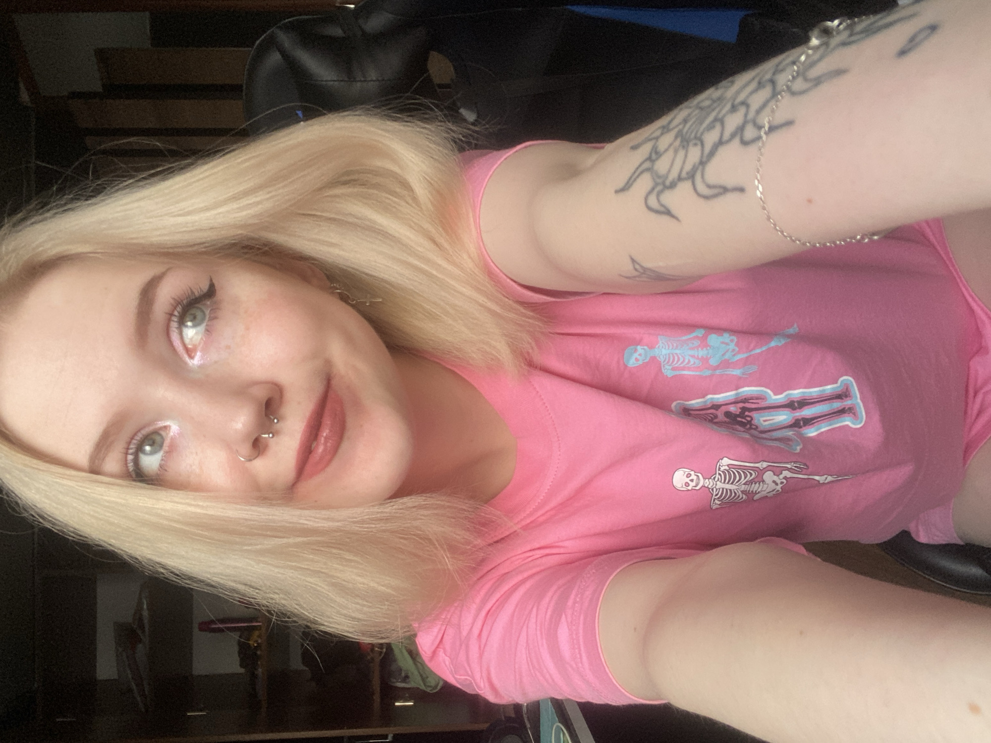 A young female with blonde hair, light skin tone, and visible freckles is depicted indoors, likely taking a selfie. She wears a pink short-sleeved t-shirt adorned with white and blue skeleton graphics, and pink shorts are partially visible. Her eyes are light-colored, enhanced with winged eyeliner and pink eyeshadow; her lips are coated with a light pink gloss. A silver septum piercing and a silver hoop earring with a cross charm are visible. She wears a silver chain bracelet on her left forearm, which also features a dark-inked tattoo, possibly depicting an arachnid or abstract design. Another lighter tattoo is faintly visible on her upper left arm. Her gaze is directed upwards and to the right, with a slight smile.
The background on the left consists of a dark shelving unit or wardrobe containing various indistinct objects, including a pink cylindrical item. On the right, the back of a black gaming or office chair is prominent, positioned against a dark wooden or paneled wall. A blue fabric item is partially visible to the right of the chair. The lighting suggests an interior setting. The precise location within the city of Izhevsk, Russia, cannot be determined from the visual information presented in the image.