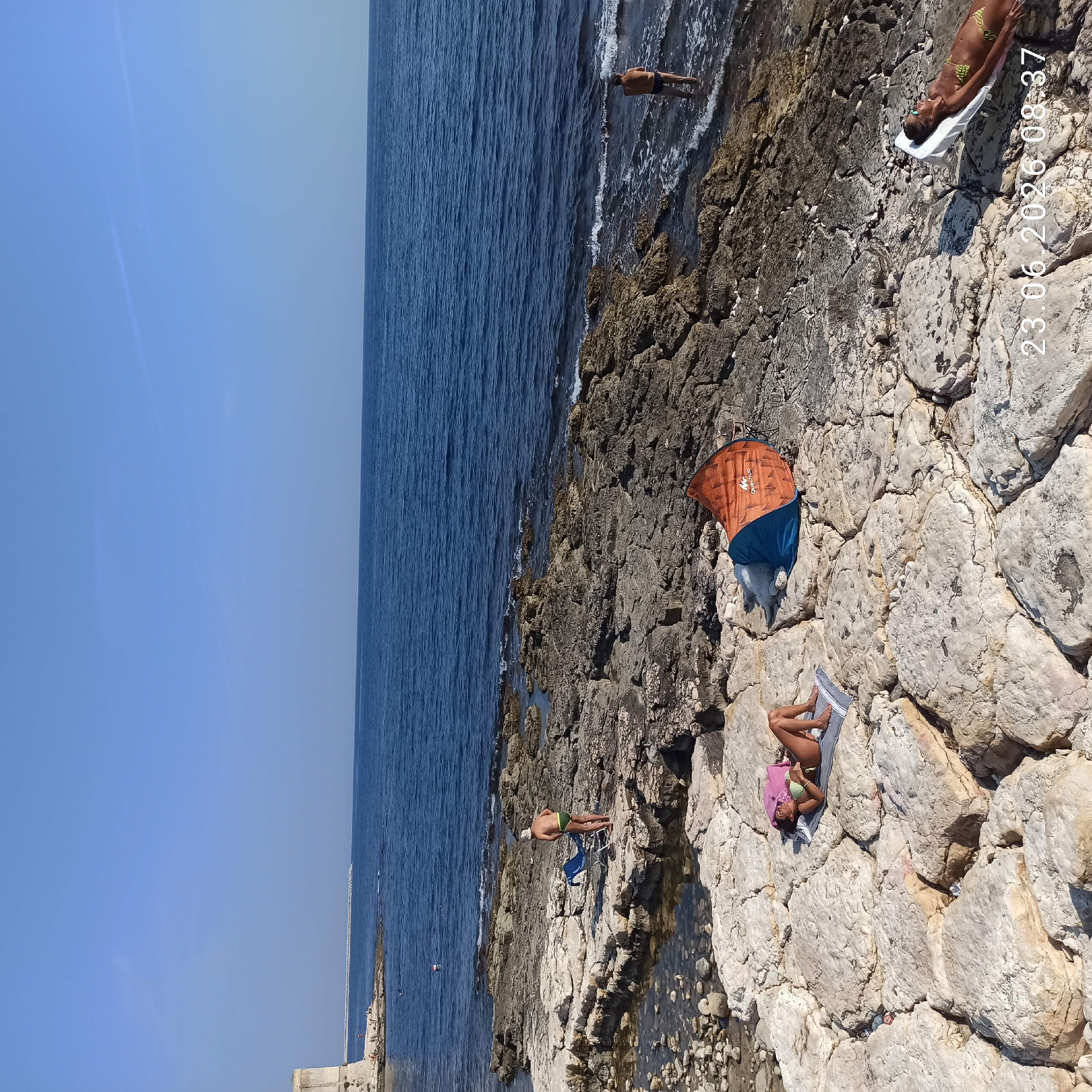 The image captures a sunny day at a rocky coastline, likely in Molfetta, Italy, given the timestamp. In the foreground, a woman in a green bikini lies on a towel on large, light-colored rocks, sunbathing. A pop-up tent, orange and blue, is set up nearby. Further back, a man in green swim trunks and a white hat stands next to a blue beach chair on similar rocky terrain. The middle ground shows more of the rocky shoreline with several people scattered around, some in the water, and a small buouy. In the distance, the deep blue sea stretches to the horizon under a clear sky. To the far left, a section of a stone structure, possibly part of a historical fortification or building, is visible at the edge of the land. The overall scene depicts a relaxed day at the beach.