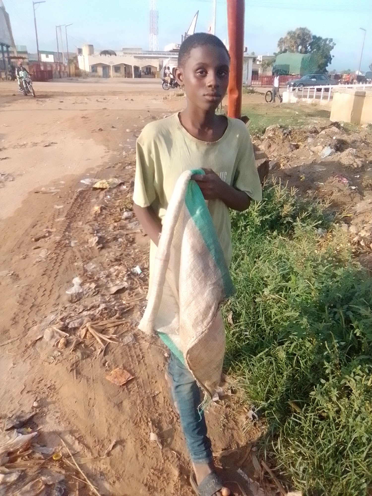 A young person stands in what appears to be a dirt lot on the outskirts of Gombe, Nigeria. The person is wearing a light-colored t-shirt and jeans, and is holding a piece of cloth with green and beige stripes. The ground is covered in dirt and debris, with patches of dry grass and green vegetation. In the background, there are buildings, a communication tower, and some vehicles, suggesting an urban or semi-urban environment. The sky is clear and the lighting indicates it is daytime.