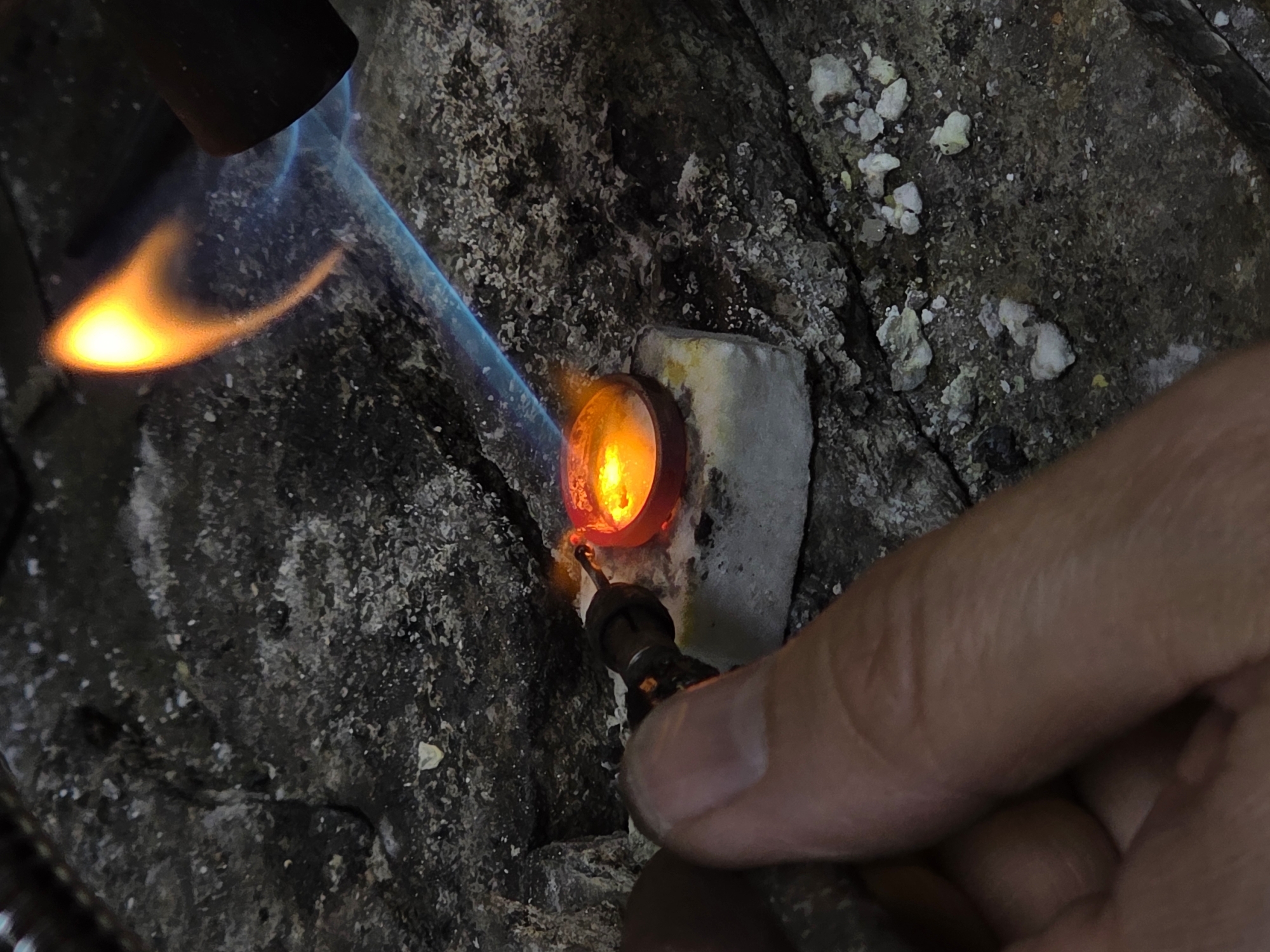 This is a close-up, vertically oriented, low-angle shot of a goldsmith at work in Kazan, Russia. The scene is dominated by a torch flame, which is bright orange and yellow at its core and transitions to blue at the edges, directed at a small, red, ring-shaped object. The object is resting on a white, porous material, likely a soldering block or crucible, which sits on a dark, rough, textured surface. A human hand, with visible finger lines and skin texture, is holding a tool that is positioned near the ring, obscuring part of the view. The background is dark and grainy, suggesting an indoor workshop environment. The overall impression is one of intense heat and focused craftsmanship. The location in Kazan is not visually discernible from the image itself.