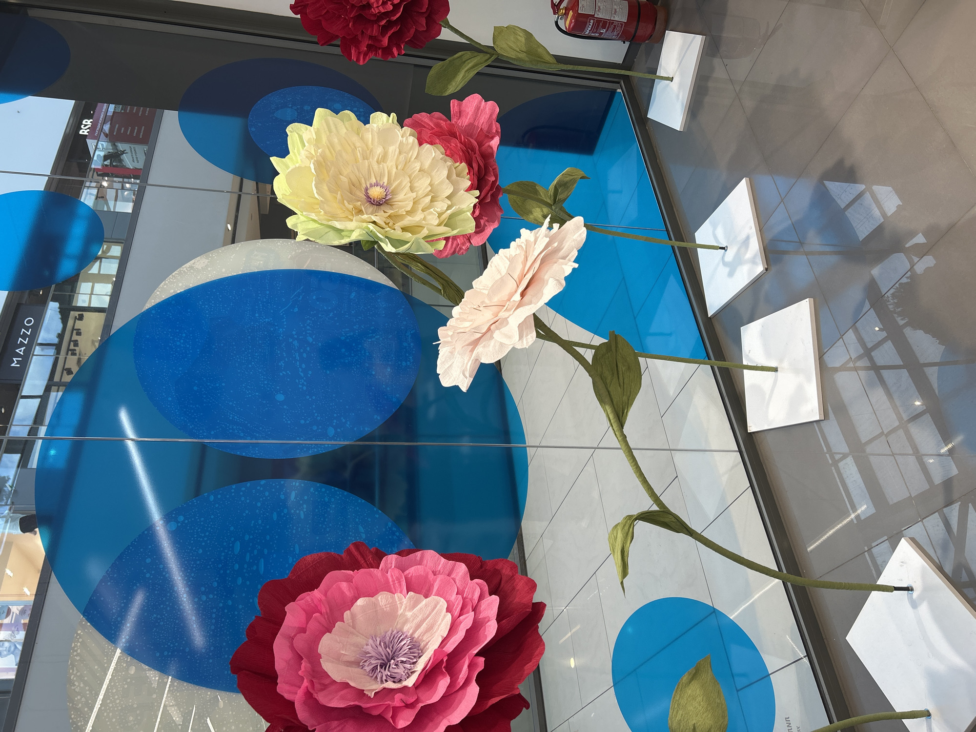 A collection of oversized paper flowers decorates a glass facade, likely a shop window, in an indoor space. The flowers are in various colors, including red, pink, and yellow, with green stems and leaves. Behind the flowers, the glass facade is adorned with large blue and white polka-dot-like circles. The word "MAZZO" is visible on the left side of the glass. The background shows reflections of what appears to be the inside of a shopping mall. An object resembling a fire extinguisher is visible at the top right. Based on the presence of the word "MAZZO," this is likely located in Sofia, Bulgaria. The flowers are fixed to the floor with white square bases. The floor tiles are reflective.