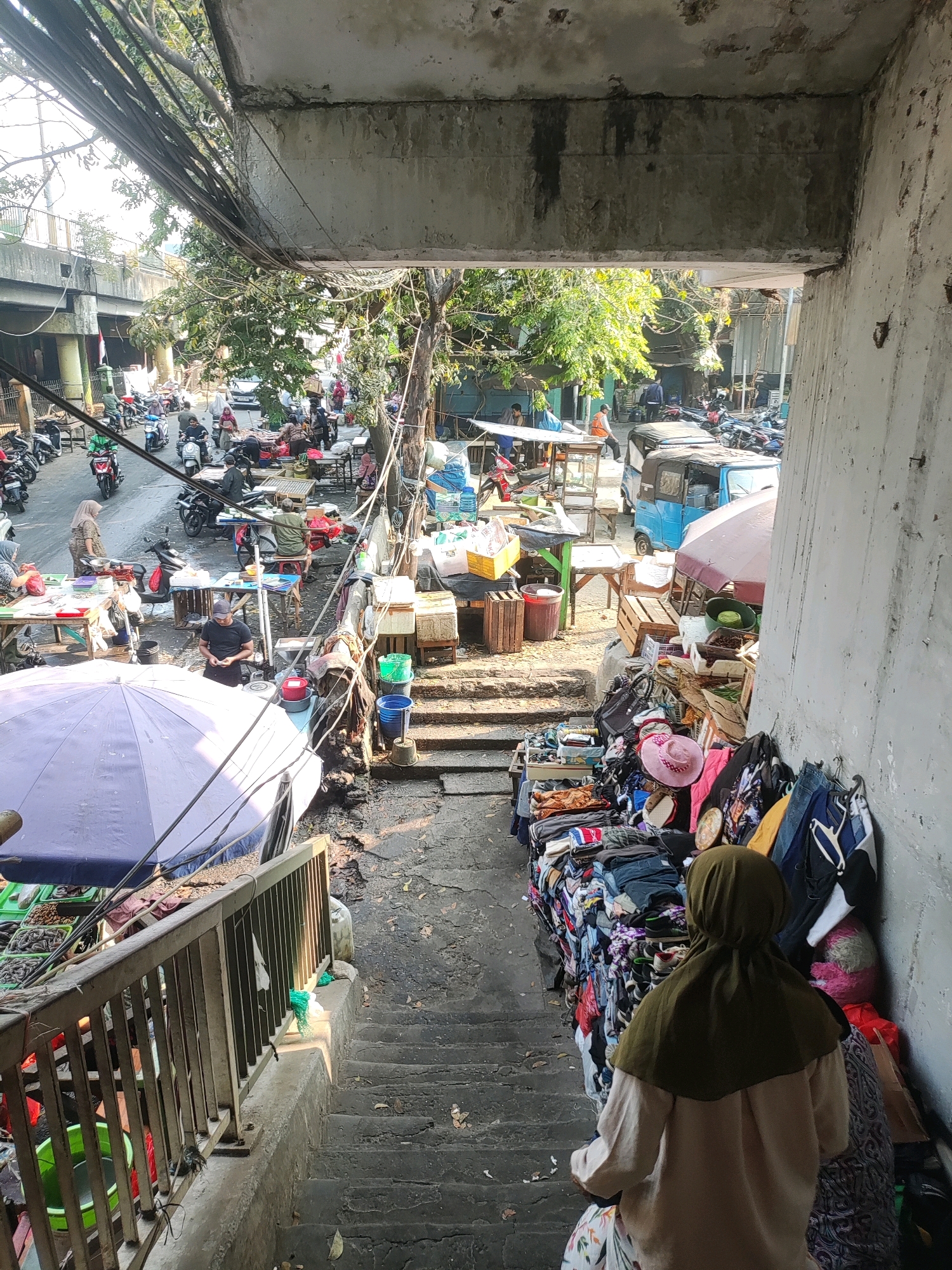 This image captures a bustling street market scene in East Jakarta, Indonesia, likely under an overpass. The foreground features a set of concrete stairs descending into the market. On the right side of the stairs, a stall is overflowing with clothing, including folded garments, hanging items, and a pink hat. A person wearing a green hijab stands at the bottom of the stairs, facing away from the camera, looking towards the market.
The market itself is filled with various stalls and vendors. To the left of the stairs, a large blue umbrella covers a stall selling what appear to be fresh produce or seafood. Beyond this, a busy street is visible with numerous motorcycles parked and in motion, and people walking and interacting. Several vendors are seen behind tables laden with goods, and some are engaging with customers. In the background, there are more stalls, some with blue awnings, and glimpses of trees and buildings. The environment appears somewhat disorganized, with overhead wires and a weathered concrete structure suggesting it's located beneath an elevated road or bridge. The overall atmosphere is one of lively commercial activity in a dense urban setting.