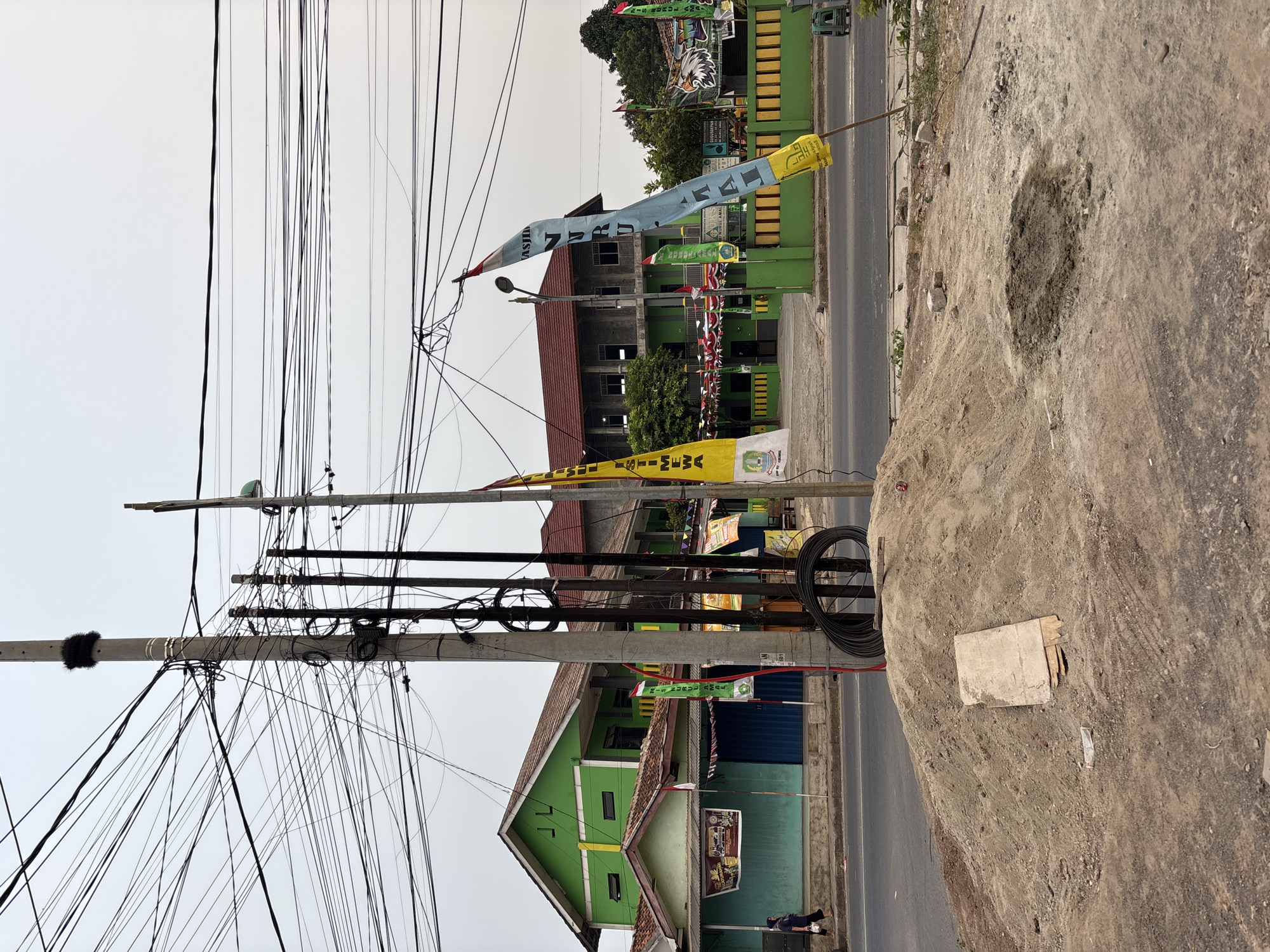 The image captures a street scene in Bekasi, Indonesia, characterized by a complex overhead network of utility wires. In the foreground, a pile of sand and construction debris sits on the side of a road. A light blue flag with yellow lettering and a red and white striped flag are planted in the sand, partially obscuring a black coil of cable. Behind the sand pile, a two-story building painted green with a red roof stands on the opposite side of the road. A smaller, light blue building with a green roof is attached to the left side of the green building, and it features closed blue roll-up doors. Two women are walking on the sidewalk in front of these buildings. Further down the road, a larger, concrete building with a red roof is visible, adjacent to the green structure. This building appears to be a school or institutional building, with green fencing and a yellow tiled wall. Another set of flags, including yellow and blue ones, are positioned in front of this building, and a mural of an eagle is present on the right side of the fence. The sky is overcast, suggesting a cloudy day. The overall impression is one of a developing or undergoing renovation area within a busy urban environment.