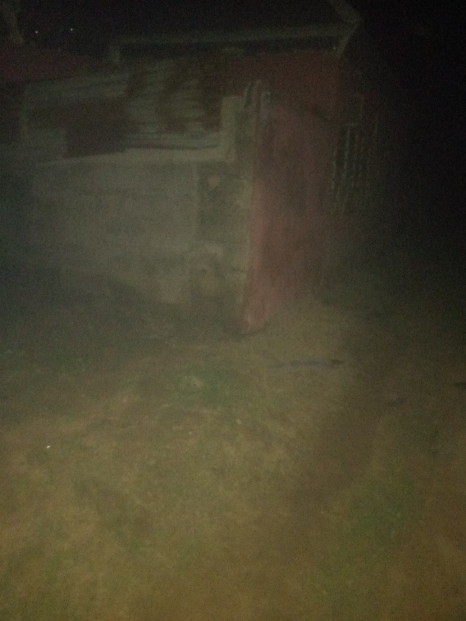 An outdoor, low-light, blurry image shows a partially constructed or dilapidated structure. The structure appears to be made of wood and possibly red-painted siding. There is a dark, possibly grassy or dirt area in the foreground, and the background is mostly dark and indistinct. The scene has a general sense of disarray.