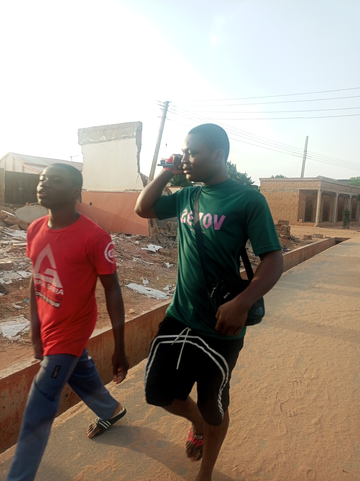 The image shows two young men walking along a paved street in Tempure, Nigeria. The man on the left is wearing a red t-shirt and blue pants, with Adidas sandals. The man on the right is wearing a green t-shirt, black shorts with white stripes, and red and black sandals. He has a black messenger bag slung over his shoulder and is holding a cell phone to his ear. In the background, there is construction debris on the left and unfinished brick structures on the right. Utility poles and power lines are visible overhead. The sky is bright with limited cloud coverage.
