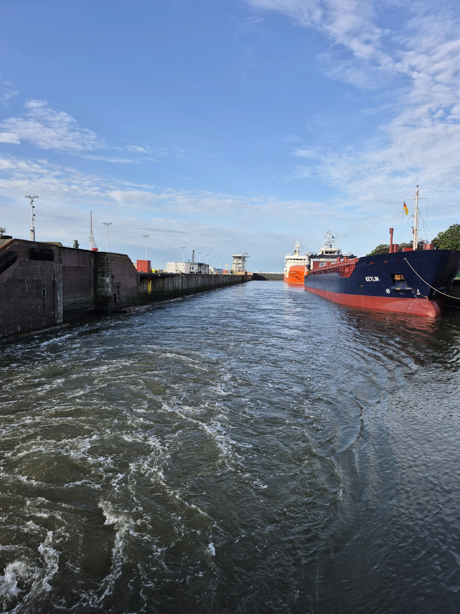 The image captures a view from a canal leading into a lock system in Brunsbüttel, Germany. In the foreground, the turbulent water suggests movement, possibly from the lock operations or passing vessels. Two large cargo ships are present within the lock. The ship on the right is dark blue with a red hull and the name "KETLIN" is visible on its side. It is docked. Another cargo ship, painted orange, is positioned behind the Ketlin. The background shows the brick structure of the lock gates on the left, and industrial infrastructure including containers, buildings, and cranes on the right bank. The sky is bright blue with scattered white clouds.