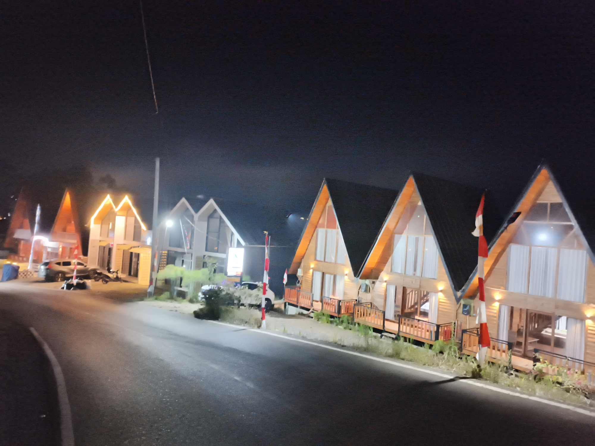 The image depicts a row of A-frame cabins at night in Dieng Kulon, Indonesia. A paved road curves in the foreground, with a white line marking its edge. On the right side of the road, several wooden cabins are illuminated from within, their triangular roofs prominently featured. Each cabin has a wooden deck with railings and large windows. An Indonesian flag is visible on a pole between two of the cabins. On the left side of the road, more cabins are visible, some with illuminated windows, and parked vehicles including a car and a motorcycle. The sky is dark, indicating nighttime. The overall impression is of a tourist accommodation area situated along a road.