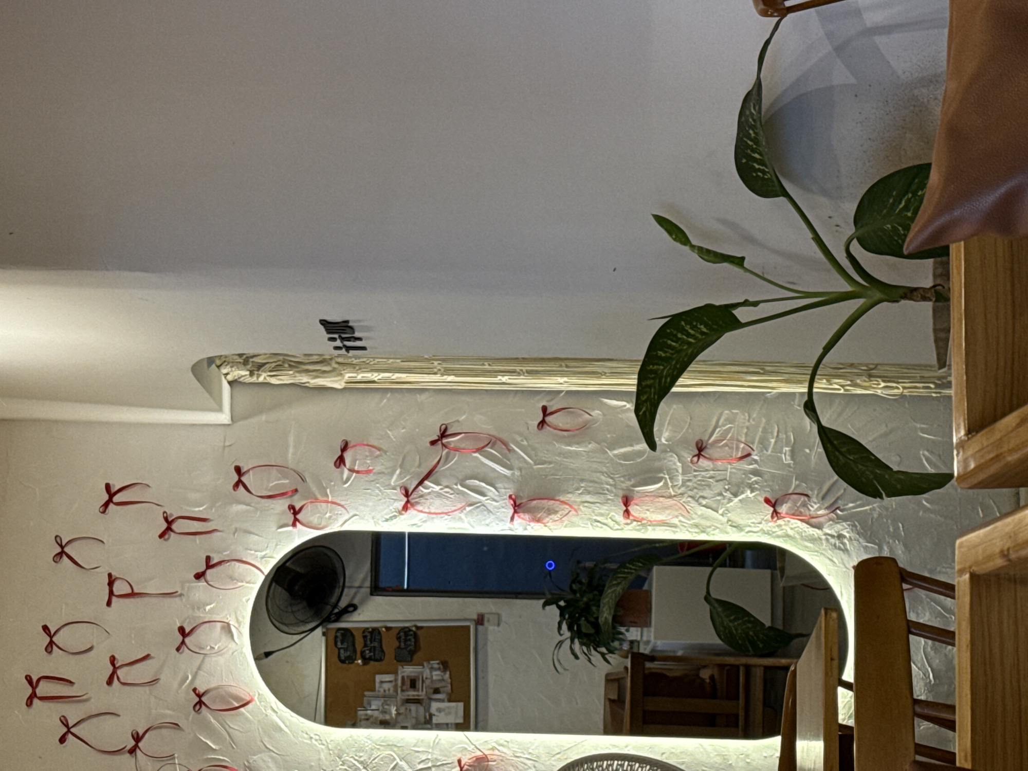 A mirror hangs on a white wall decorated with red ribbon fish shapes. The mirror reflects a fan, a corkboard with items pinned to it, a plant, and part of the room. A plant and a wooden structure are visible on the right side of the frame. The image appears to be taken in a home, likely in Bac Nha Trang Ward, Vietnam.