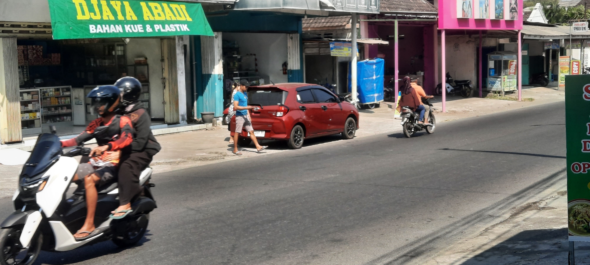 The image depicts a street scene in Selopuro, Indonesia, with a focus on pedestrian and vehicular activity. In the foreground, two individuals are riding a white and black scooter, both wearing helmets. The rider in front has a dark hooded jacket and the passenger behind is wearing an orange shirt. To their left, a red car is parked on the side of the road. Two pedestrians are standing near the car, one in a blue t-shirt and shorts, and another in a patterned dress and headscarf. Further down the street, another motorcycle with two occupants, a man in an orange shirt and a woman with a headscarf, is moving away from the camera. The environment features buildings with storefronts, including one with a green banner that reads "DJAYA ABADI BAHAN KUE & PLASTIK." The road is paved, and there is a sidewalk area where the pedestrians are located. The scene appears to be set during the daytime under sunny conditions, as indicated by the shadows cast on the road.