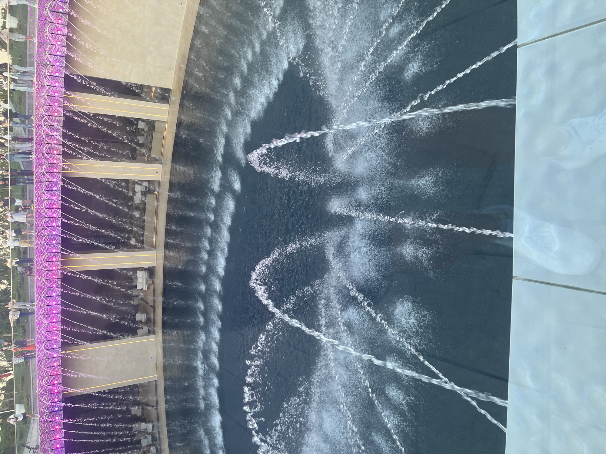 This image captures an illuminated fountain installation, likely during the evening or night, given the prominent artificial lighting. The scene is viewed from an elevated perspective, looking down into a large, dark body of water.
On the left side of the frame, a long, curved structure with a prominent row of purple LED lights lines what appears to be a walkway or viewing platform. Numerous small water jets are visible, shooting in arcs from this structure into the main fountain pool. Above this lit edge, reflections of many people can be seen in the water, indicating they are standing along the barrier, observing the fountain. The people themselves are also visible in the upper-left, appearing as a blurred crowd.
The central and right portions of the image feature the main fountain pool, filled with dark blue-grey water. Several powerful water jets are actively shooting across the pool, creating distinct, luminous streams and splashes on the surface. These larger jets form various patterns, some appearing to crisscross, while smaller ripples and splashes are also visible across the water's surface.
On the far right, a section of a light-colored, possibly tiled or reflective surface is visible, which seems to be part of an adjacent walkway or platform. A faint reflection of what resembles a person's footwear is discernible on this surface.
The overall atmosphere suggests a public gathering place in Krasnodar, Russia, where people are enjoying a vibrant and dynamic water display. There is no visible text in the image.
