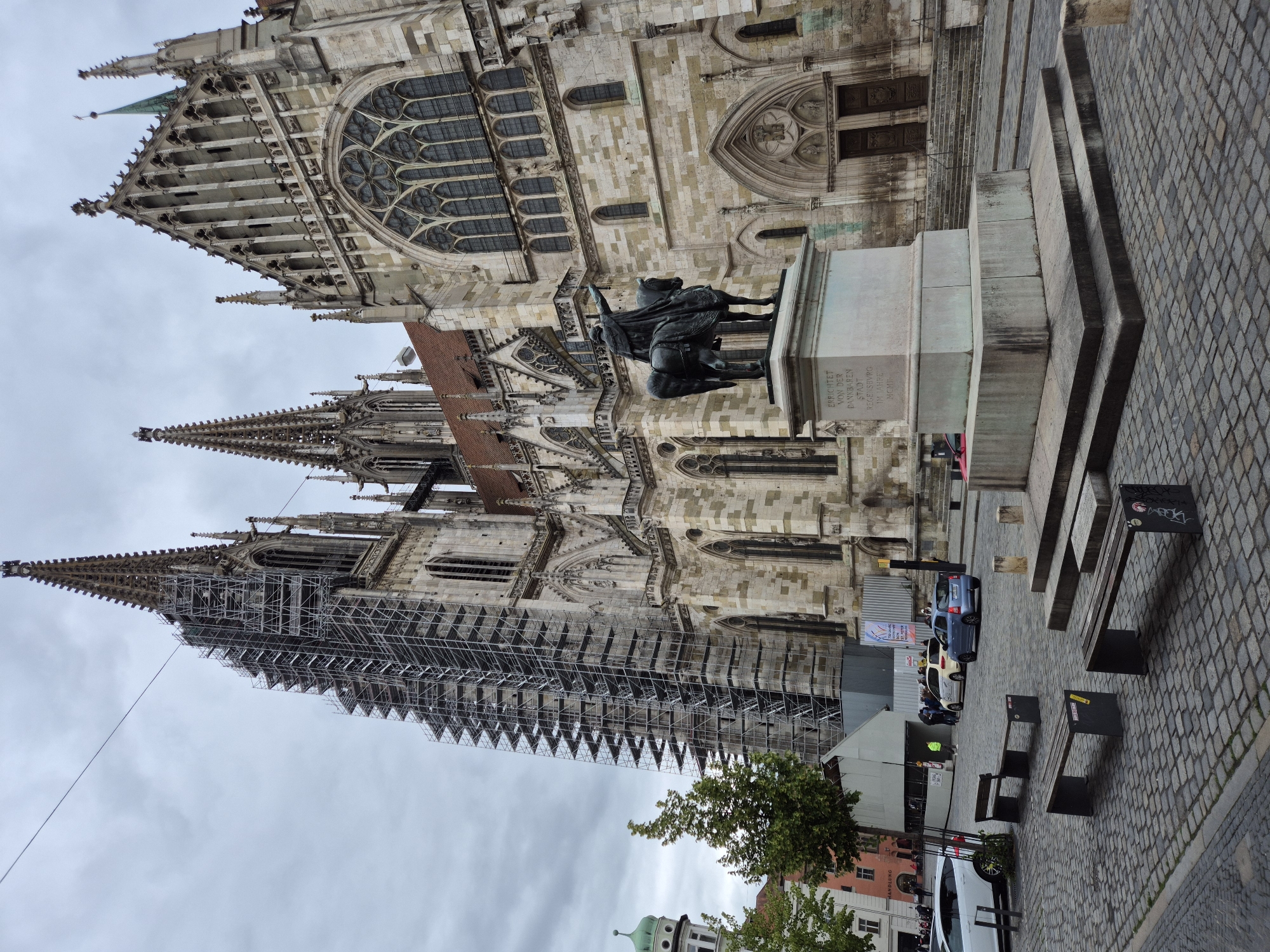 The image depicts the Regensburg Cathedral (Dom St. Peter) in Regensburg, Germany, on a cloudy day. A large portion of one of the cathedral's spires is covered in scaffolding, indicating ongoing renovation or maintenance. In the foreground, a bronze equestrian statue stands on a stone pedestal. The statue depicts a figure on horseback, with the rider pointing forward. Inscriptions are visible on the pedestal. The ground is paved with cobblestones. Several benches are positioned in front of the statue. Behind the statue and to the right, the grand facade of the Regensburg Cathedral is visible, with its intricate Gothic architecture, large arched windows, and double doors. To the left of the cathedral, modern buildings and trees can be seen, as well as parked cars and a few people. The overall scene is a mix of historical architecture and modern elements.