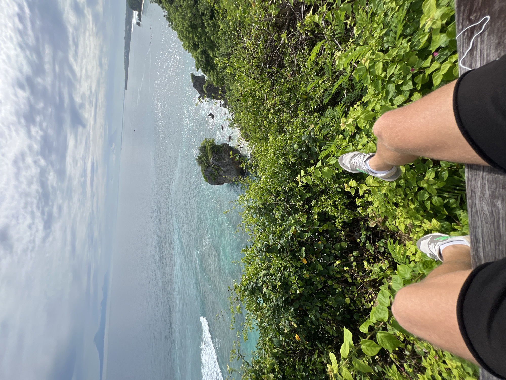 A person is seated, legs dangling, overlooking a coastline. The scene is Kuta Selatan, Indonesia. The person wears shorts and sneakers, their legs close to the edge. Below, waves crash against rocks. Dense green foliage grows right up to the edge. The ocean stretches into the distance under a cloudy sky.